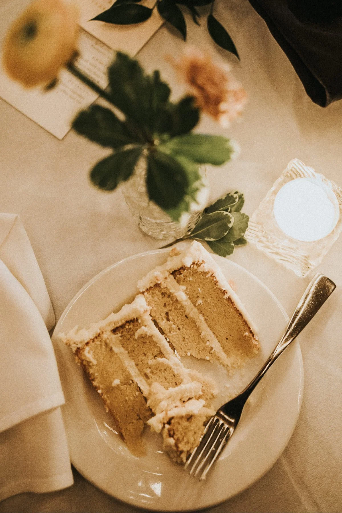 Slice of cake with fork on white plate, next to flowers and candle.