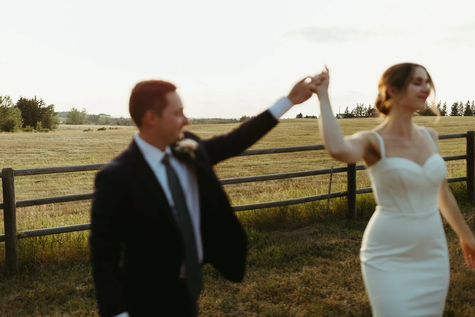Blurred image of a couple dancing outdoors in a field, with a wooden fence and distant trees in the background.