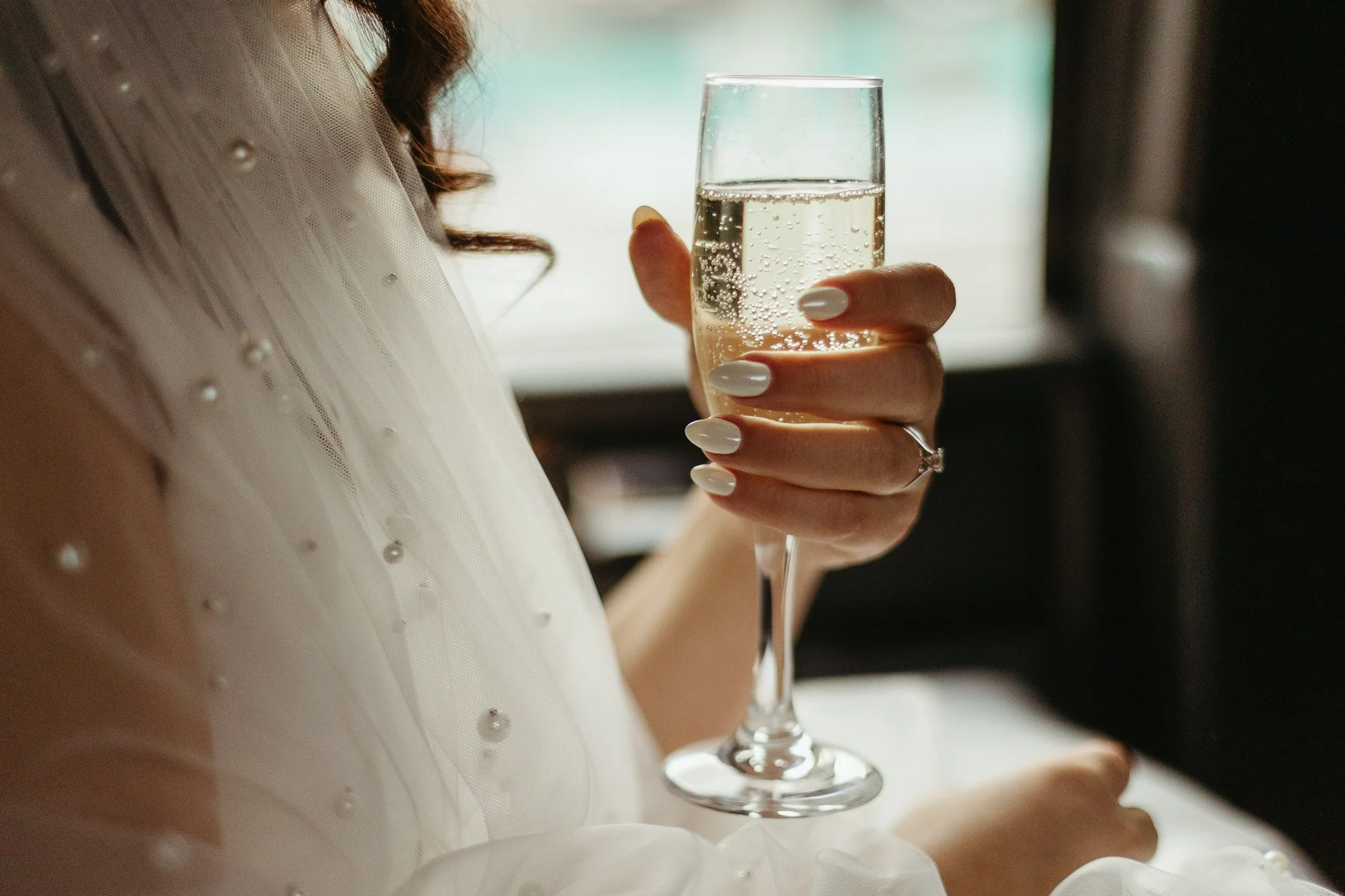 Bride with pearl nails and pearl wedding veil holds glass of champagne in the back of her wedding limo