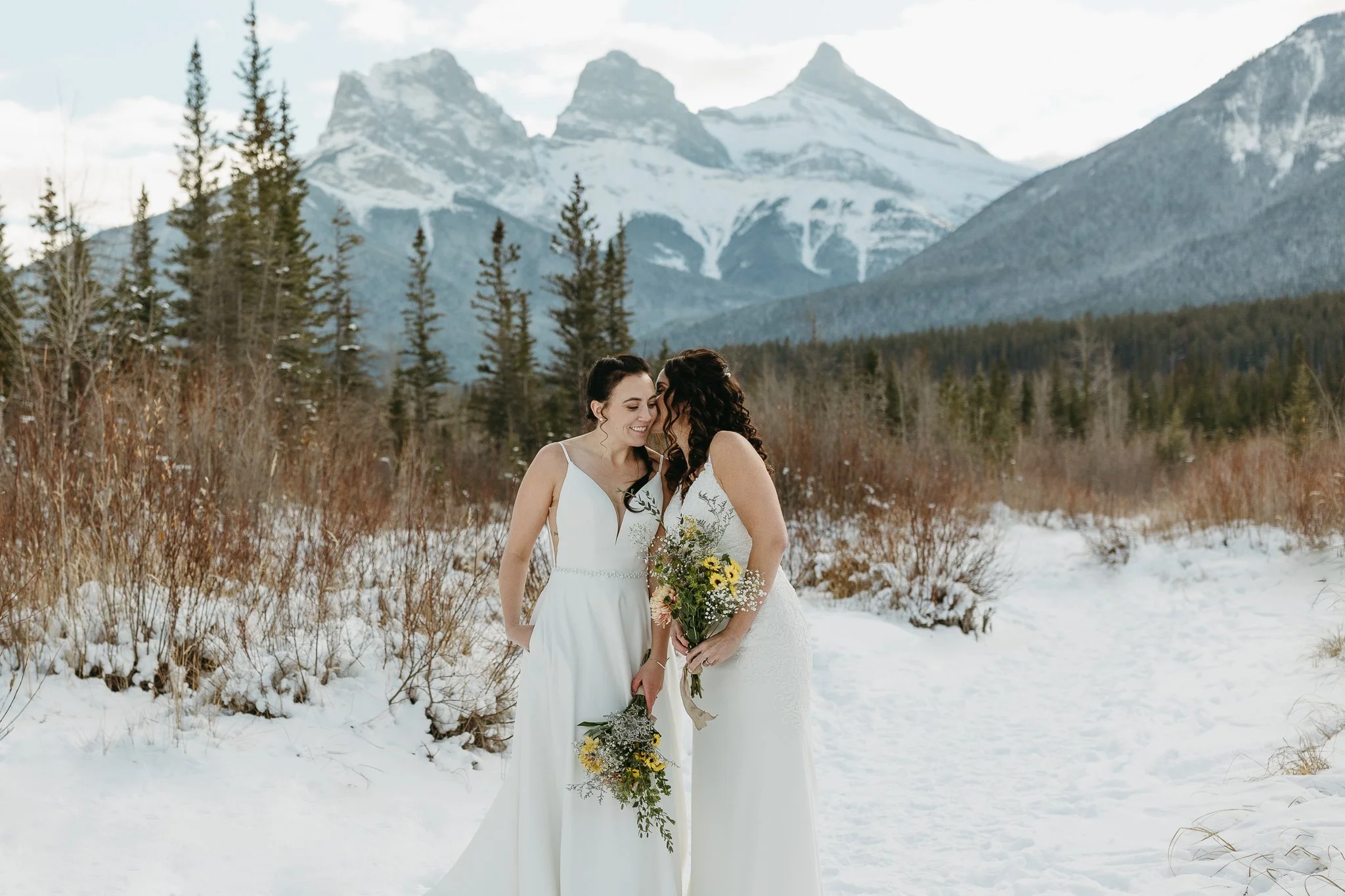 Two brides hug on their wedding day in Canmore with snowy mountains in the background