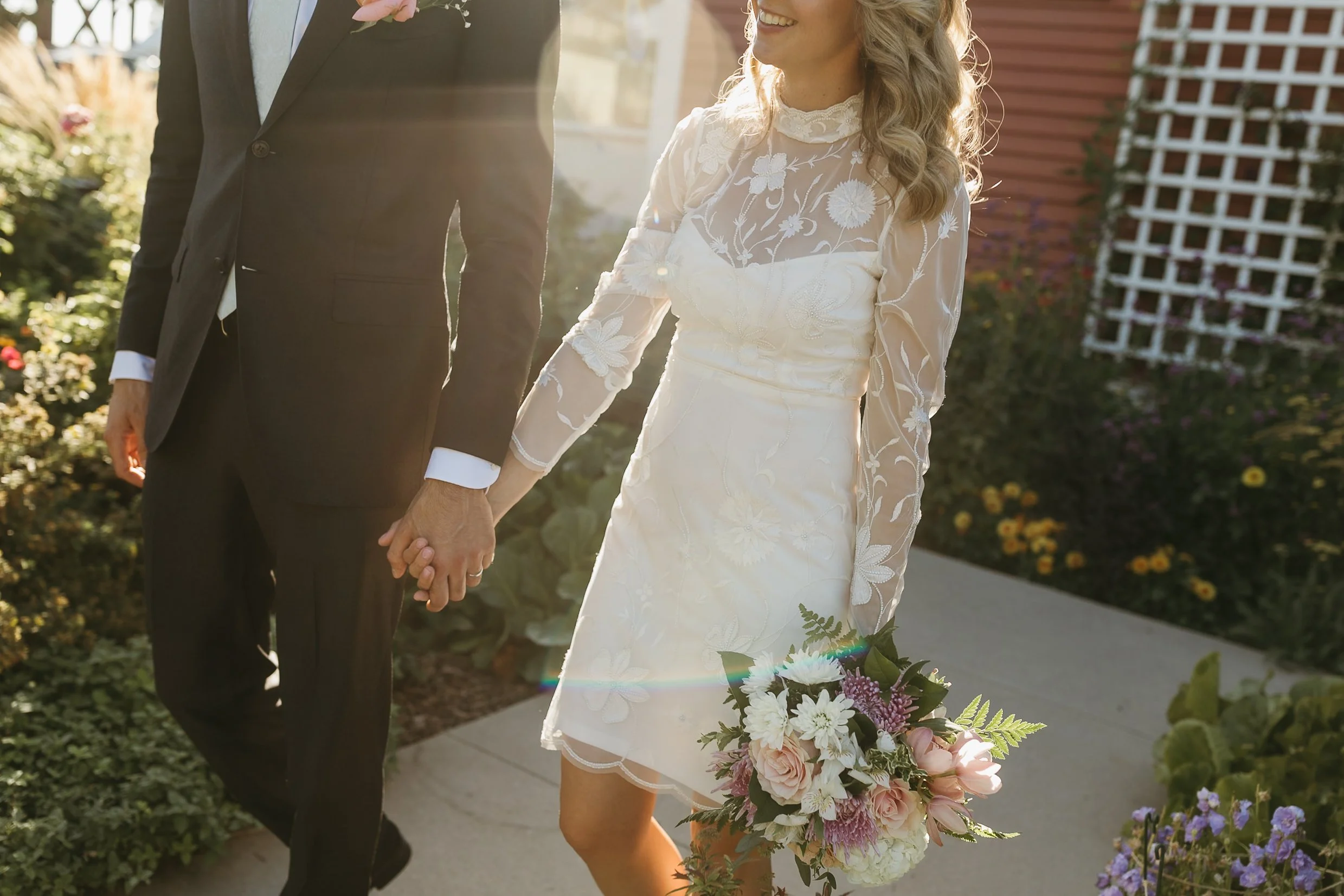 A bride and groom holding hands in the garden at Deane House on their wedding day in Calgary, with the bride holding a bouquet of flowers, during a wedding ceremony.