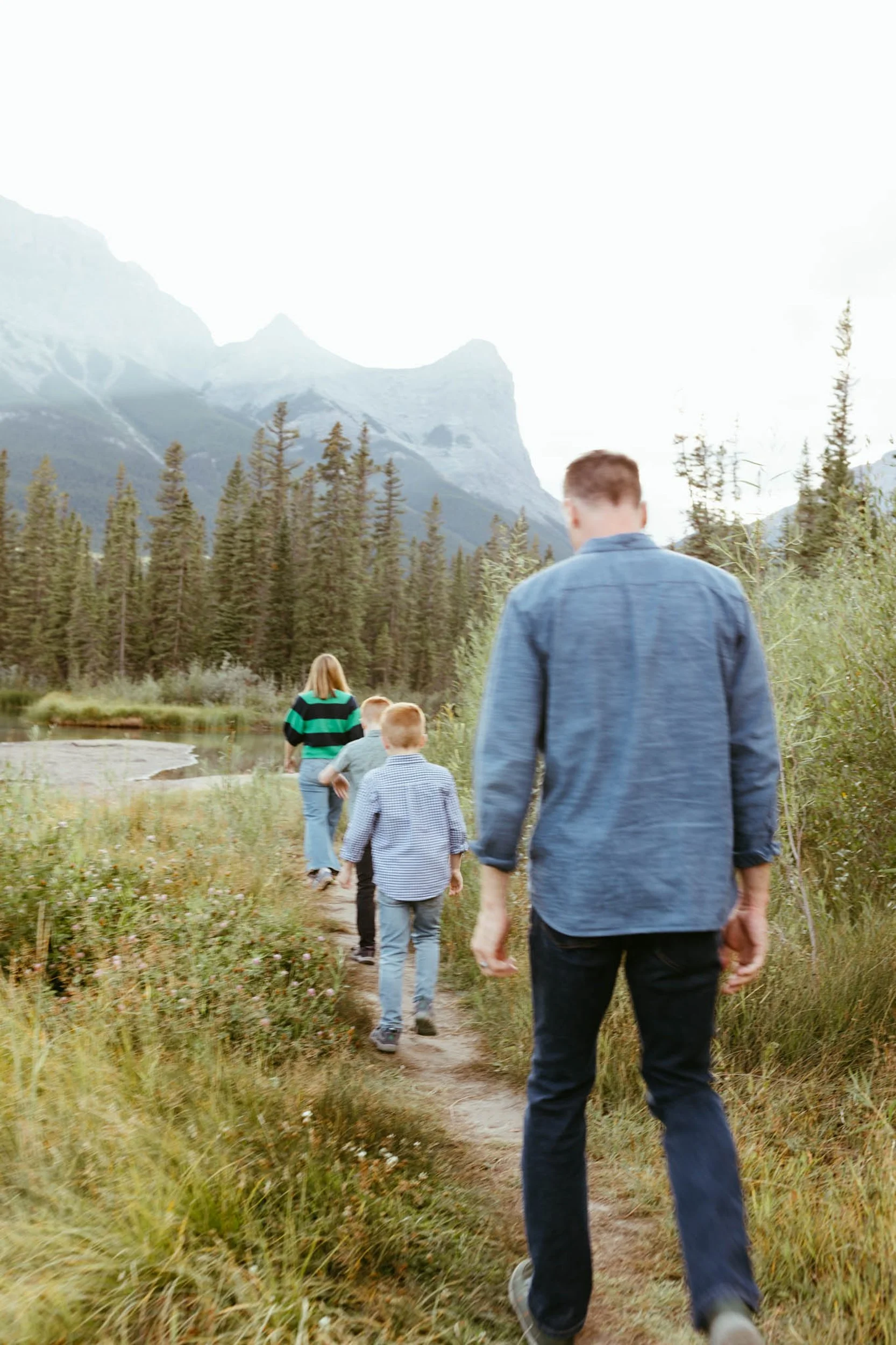 A man and children hiking on a trail in a scenic mountain area with tall trees and snow-capped peaks in the background.