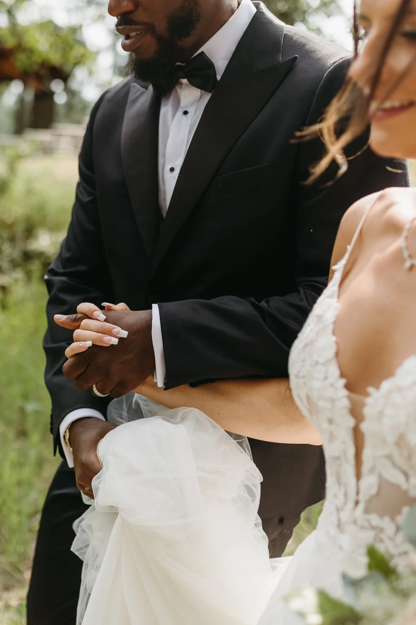 A bride and groom holding hands during their wedding ceremony outdoors. The groom is dressed in a black tuxedo with a bow tie, and the bride is wearing a lace wedding gown and veil.