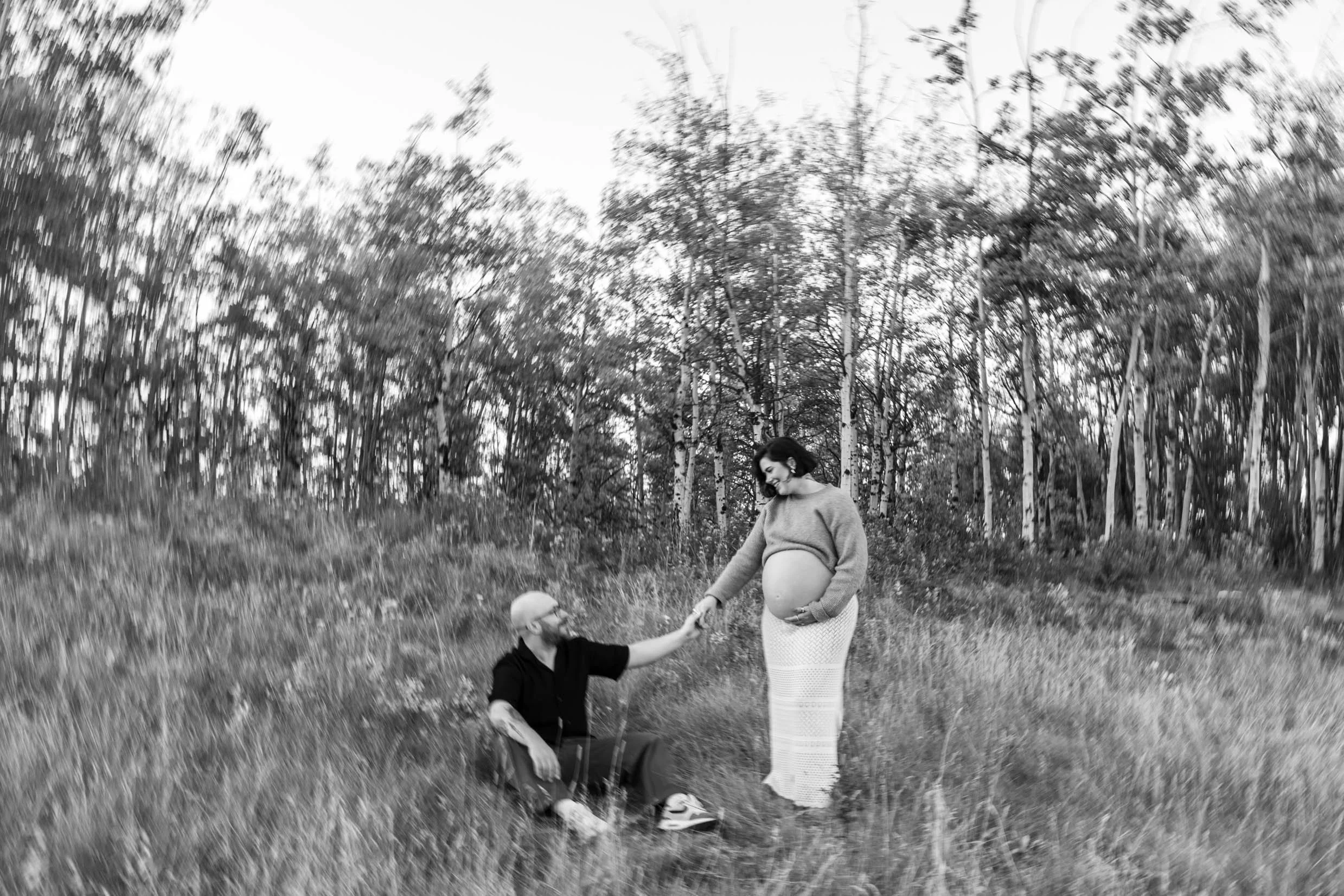 Black and white photo of pregnant woman in long skirt and sweater holding her husband's hand during fall maternity session in Calgary 