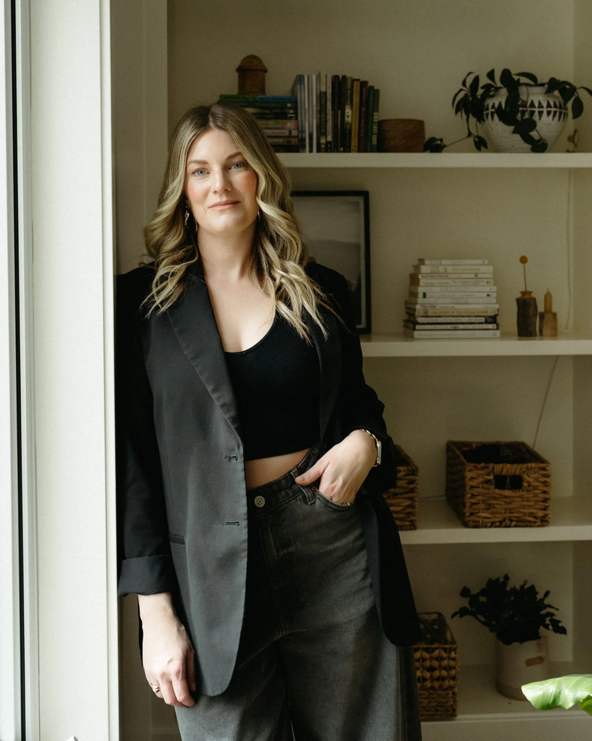 A woman with shoulder-length blonde hair, wearing a black blazer and black crop top, standing indoors near a window with a bookshelf and decorative items in the background.