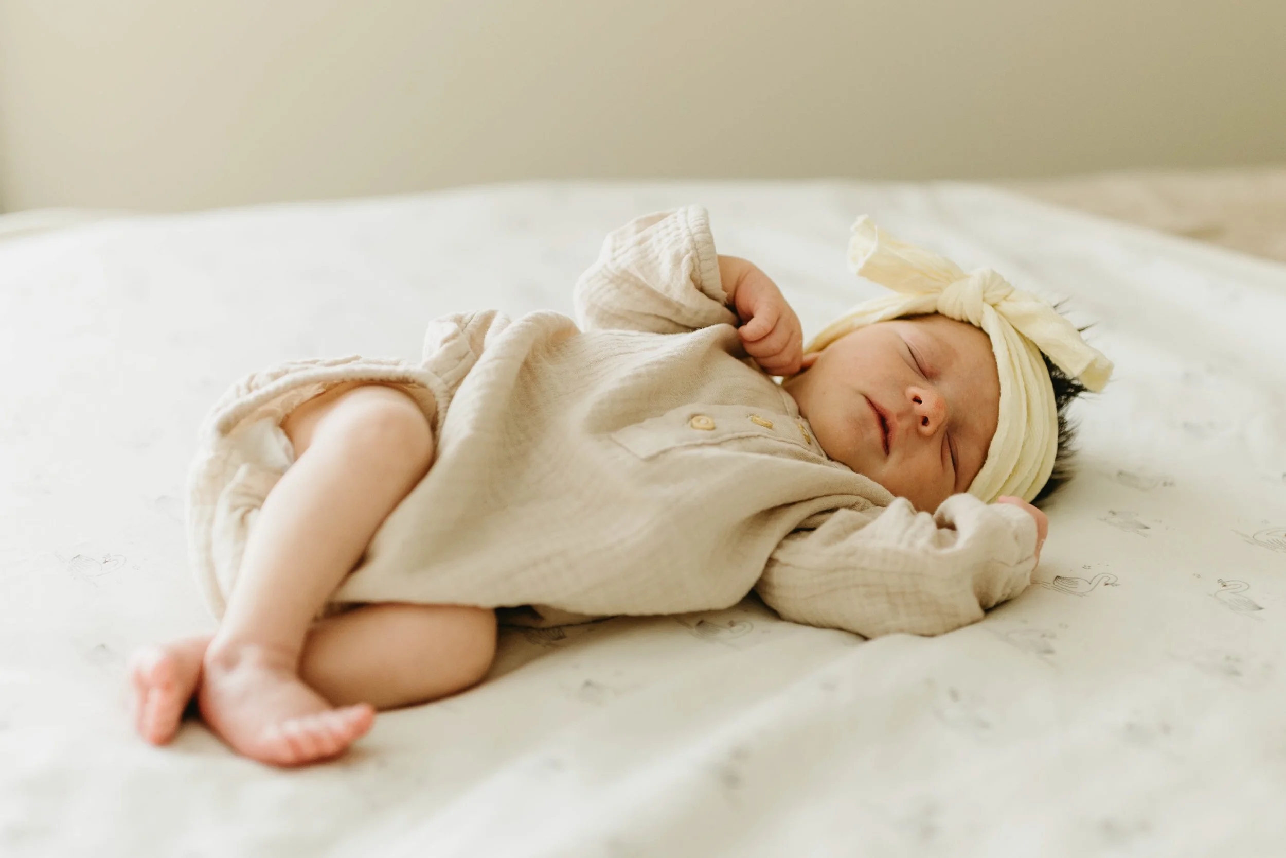 A sleeping newborn baby girl lying on a bed with a soft, neutral-colored blanket, wearing a beige outfit and a yellow headband with a bow.