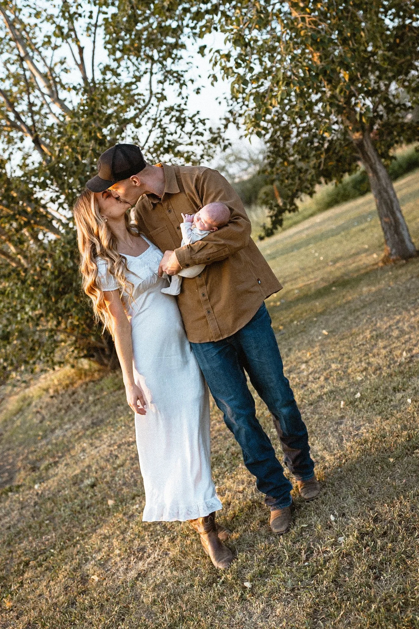 A couple, a man and a woman, sharing a kiss outdoors while holding their baby, standing in a grassy area with trees, during sunset.