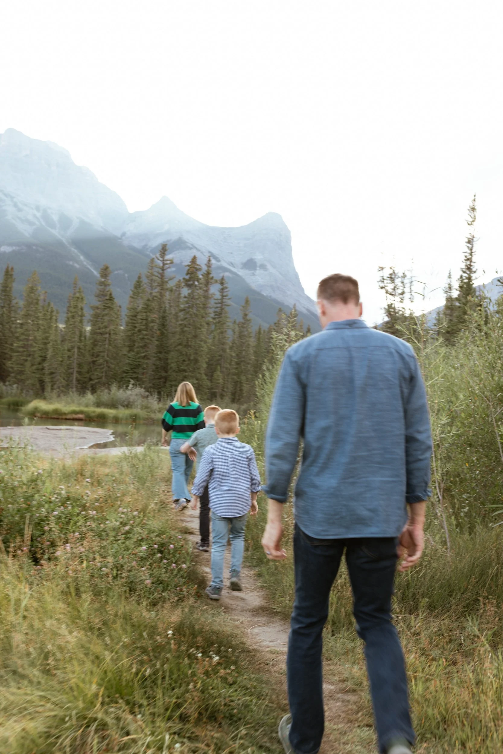 A family of five, including a man and woman with three children, walking on a trail near a lake surrounded by trees and snow-capped mountains.