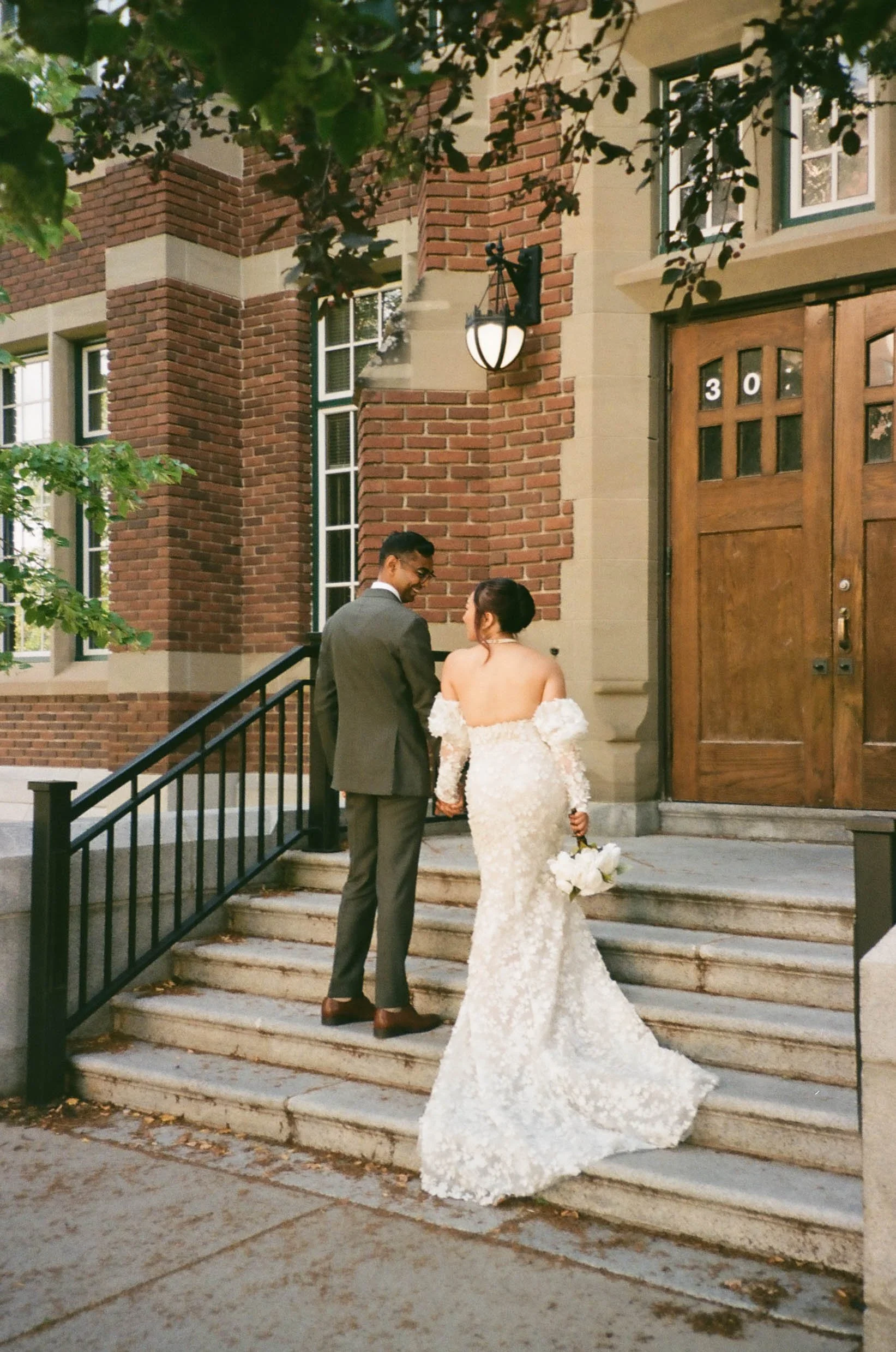 35mm film photo of groom in olive green suit and bride in ivory dress walking up the stairs with brick building in the background on their wedding day at SAIT in Calgary, Alberta