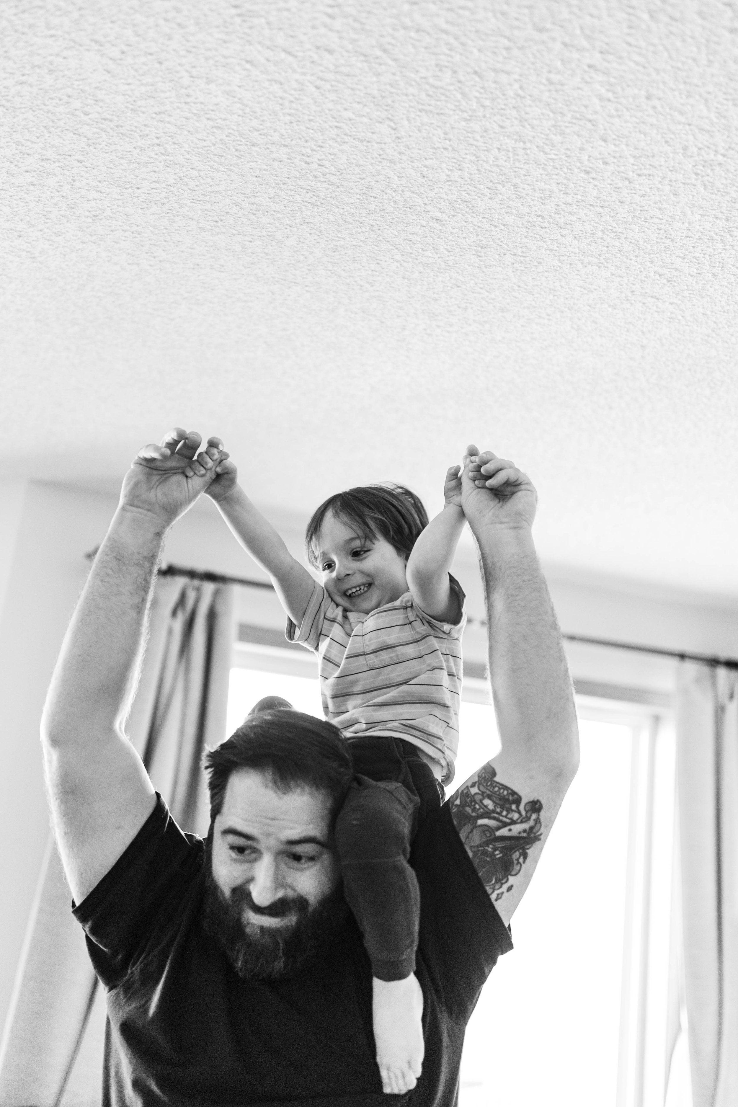 A father in a black short holds his son on his shoulders in their living room as they both laugh candidly