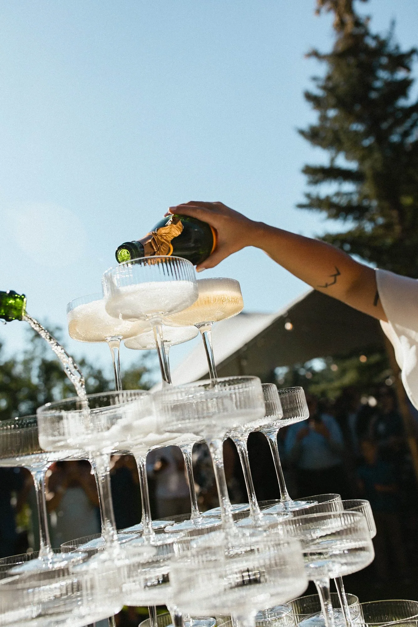 A person pouring champagne into a tower of glasses outdoors during daytime.