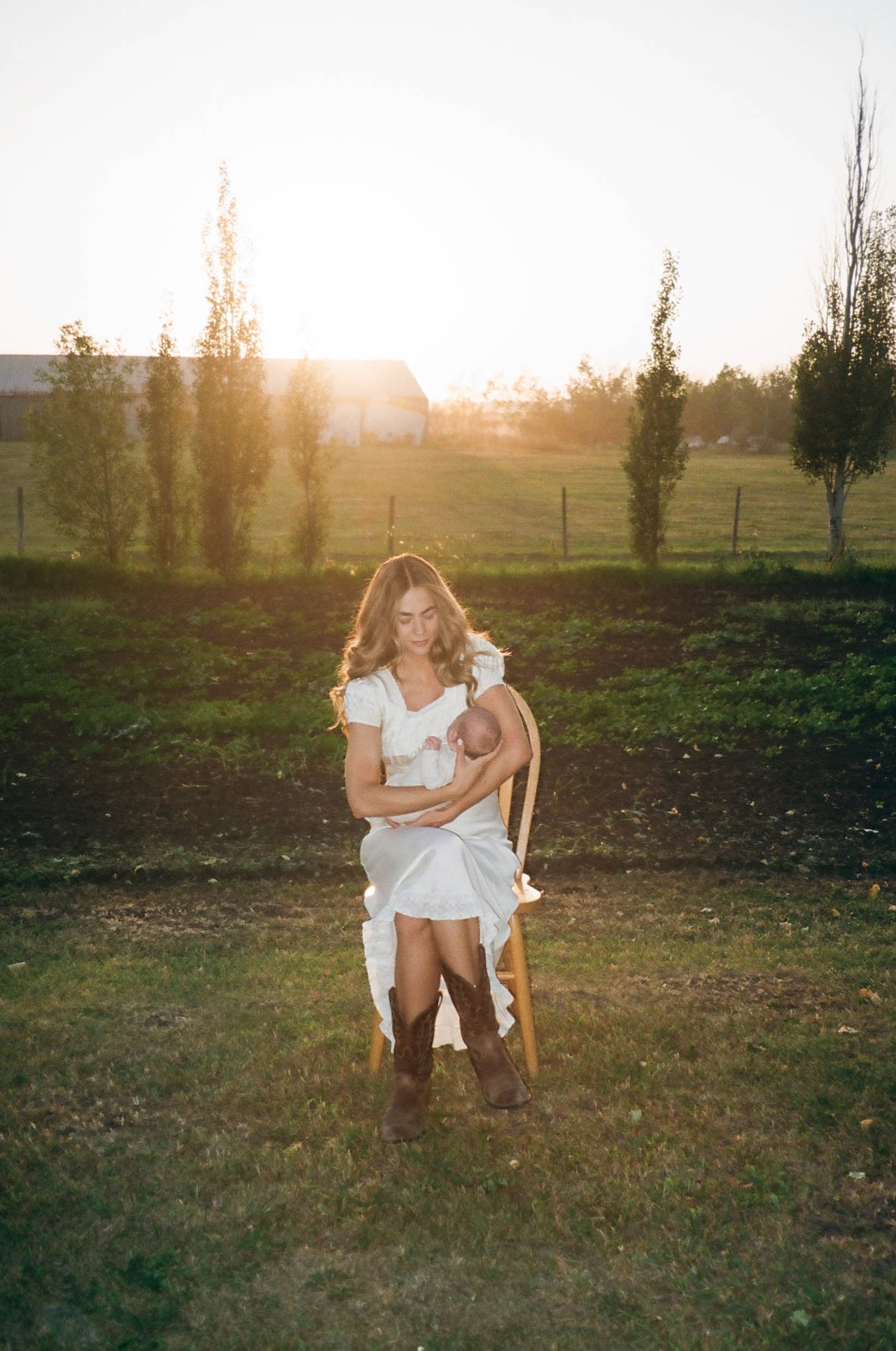 A woman in a white dress sitting on a chair outdoors during sunset, holding a baby.
