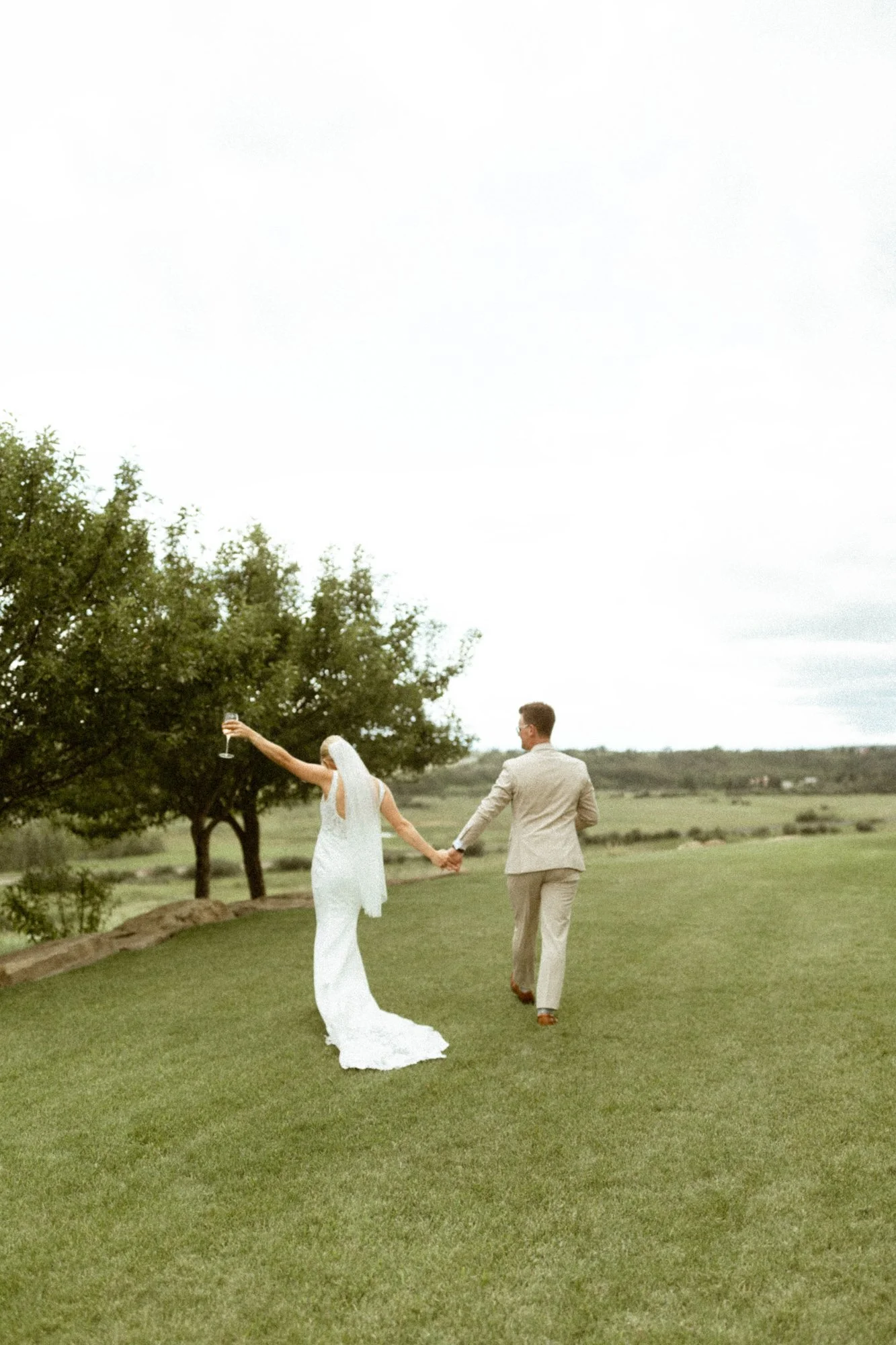 Bride and groom holding hands, walking on green grass outdoors, bride holding a glass of wine, trees and a cloudy sky in the background.