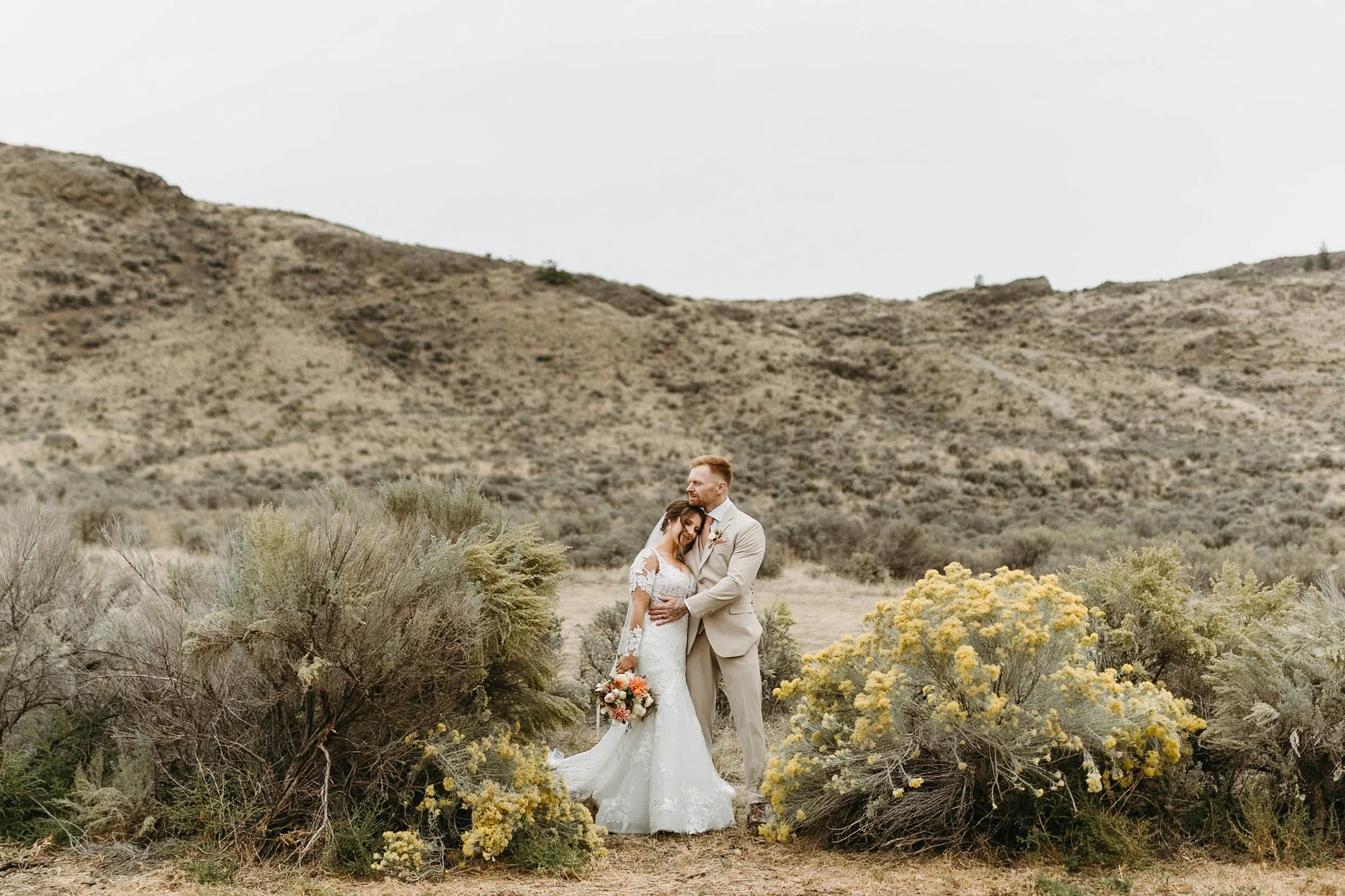 Bride in lace wedding dress with sleeves hugs groom in tan coloured suit amongst the blooming yellow sagebrush on their wedding day at Rancho Burro in Kamloops