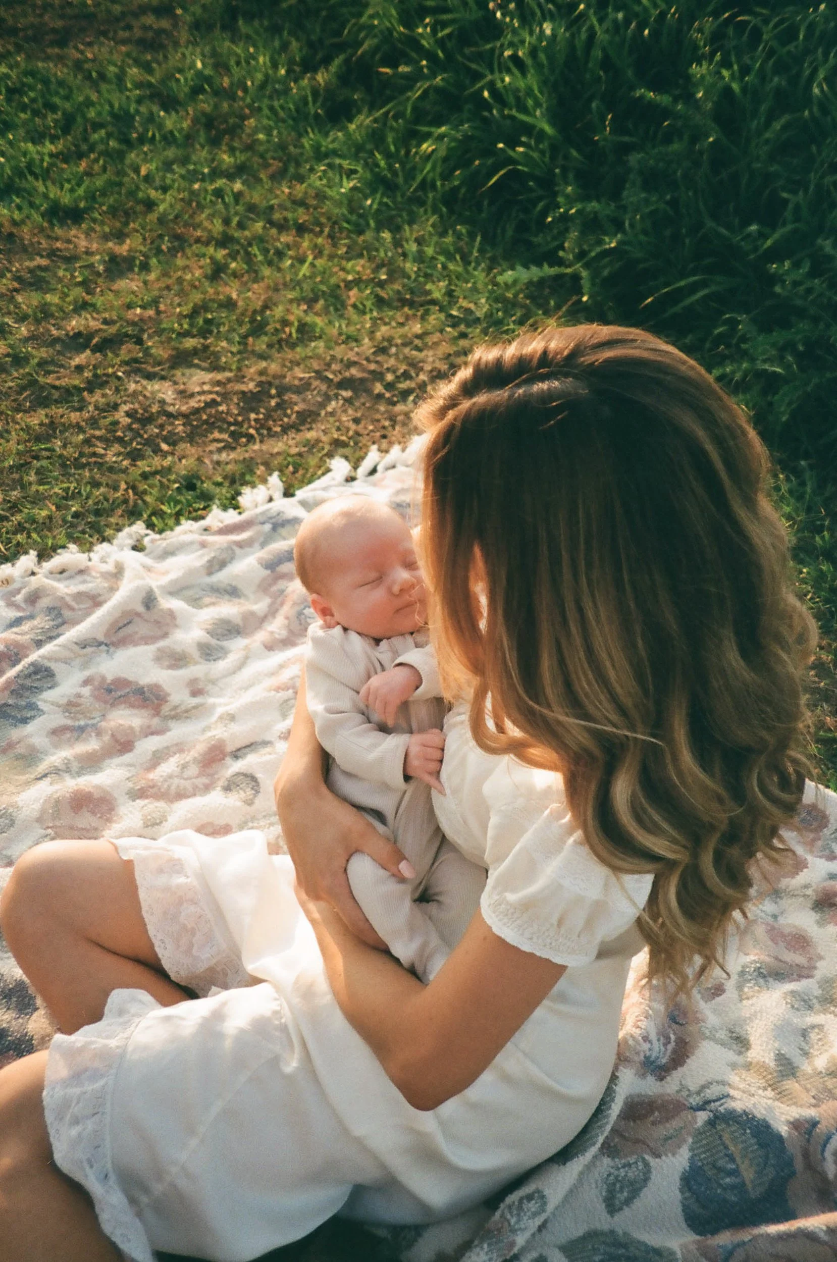 A woman holding a sleeping baby while sitting on a blanket outdoors in a grassy area during sunset.