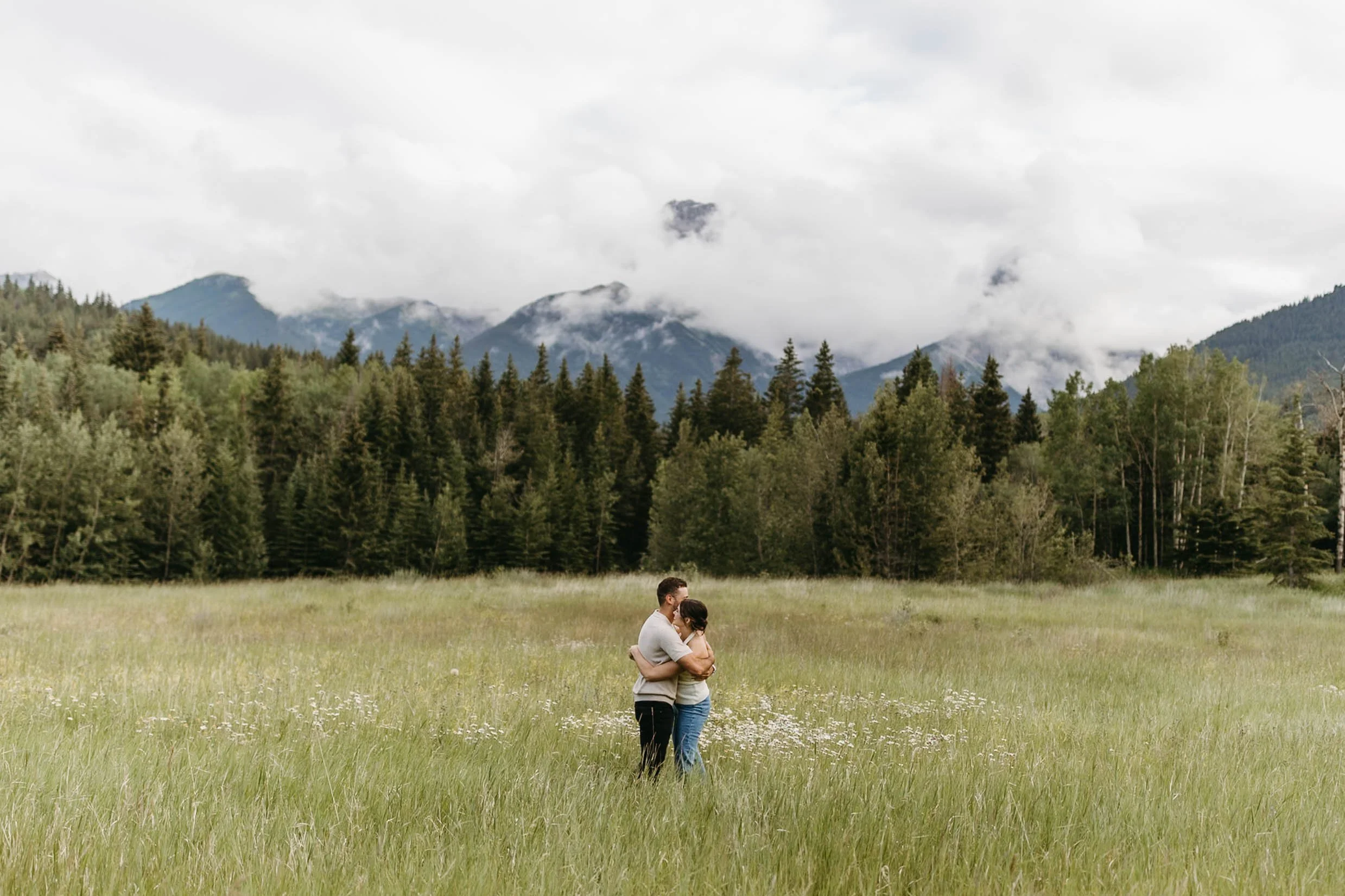 Candid photo of couple hugging in picturesque meadow with wild flowers and foggy mountain views during engagement session in Canmore