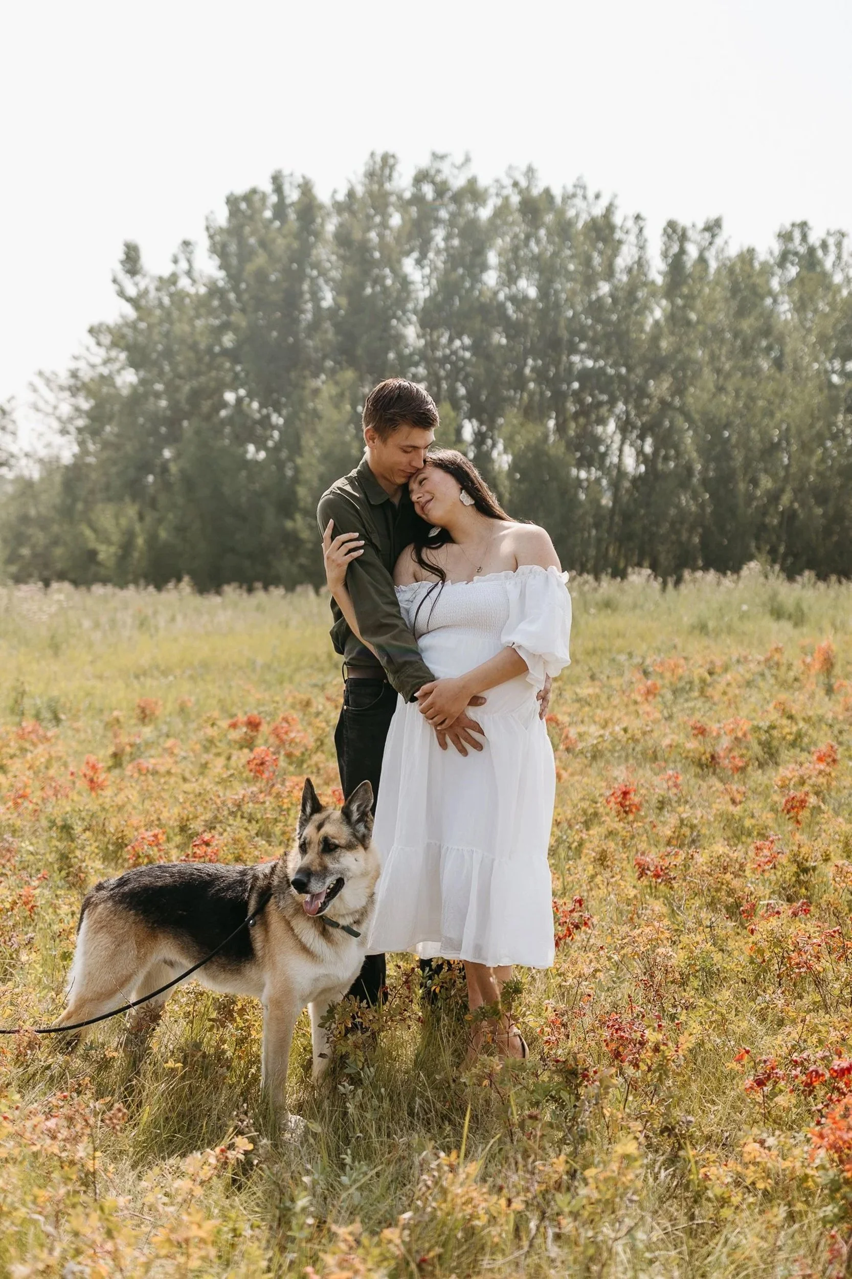 A couple sharing a kiss with a pregnant woman in a white dress, accompanied by a dog, in a field with flowers and trees in the background.