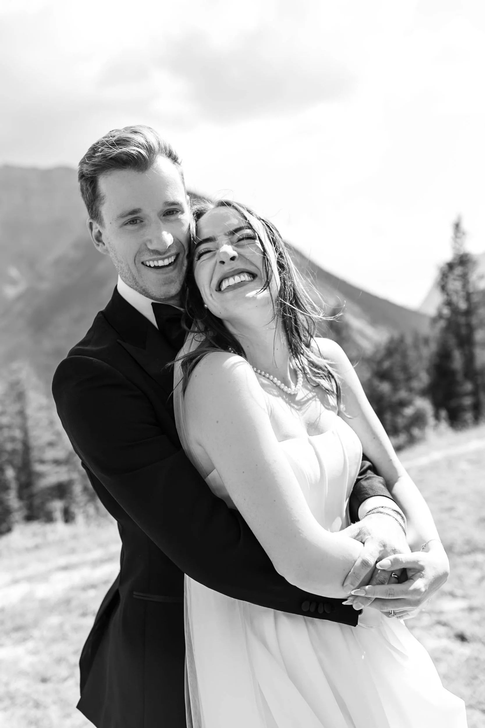 A smiling couple dressed in wedding attire, embracing outdoors with mountains and trees in the background.