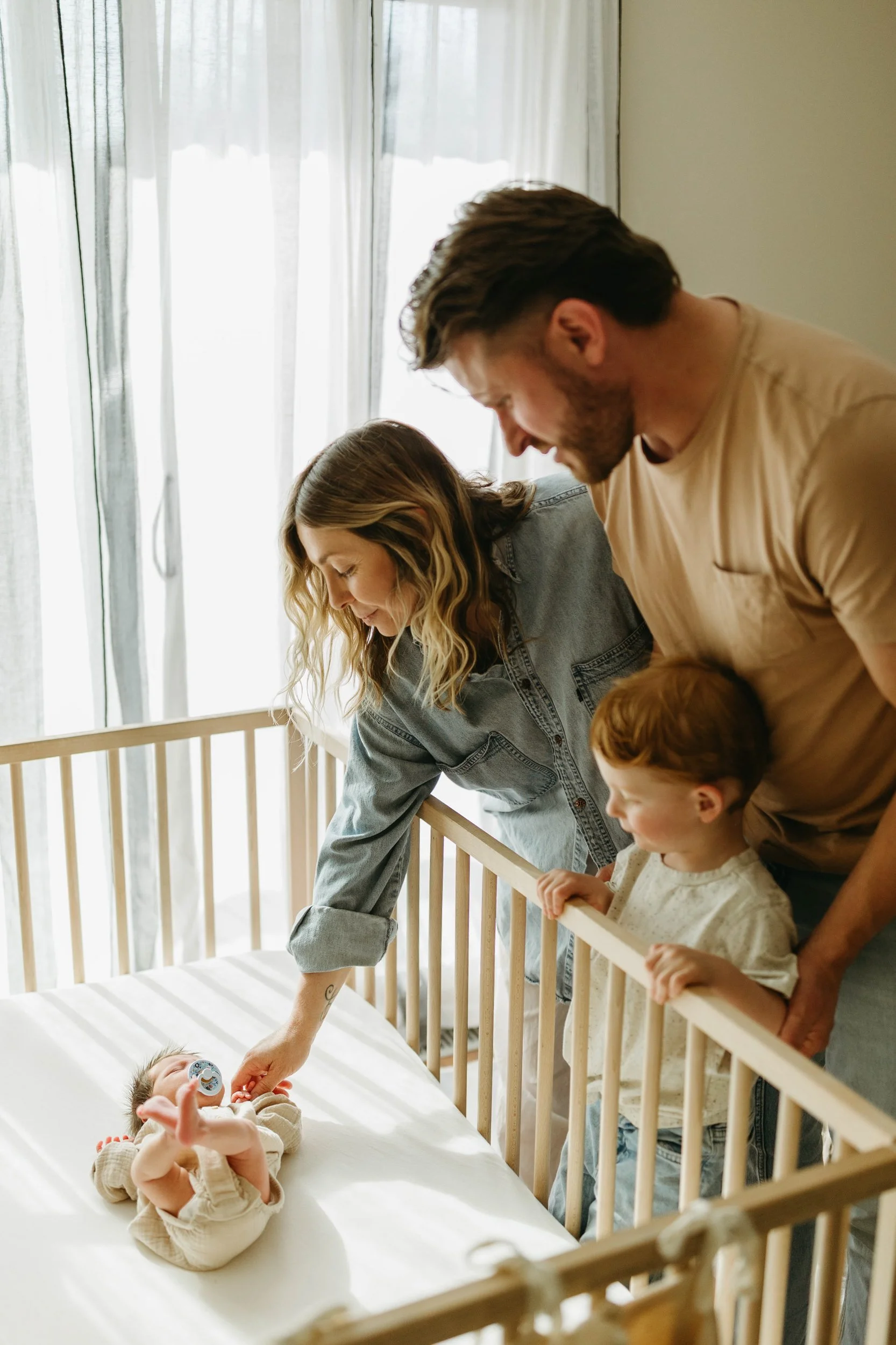 A family of four, including a mother, father, young boy, and baby, gathered around a crib in a sunlit room. The mother is gently touching the baby who is lying in the crib. The family appears to be caring for the baby in a cozy, bright setting.