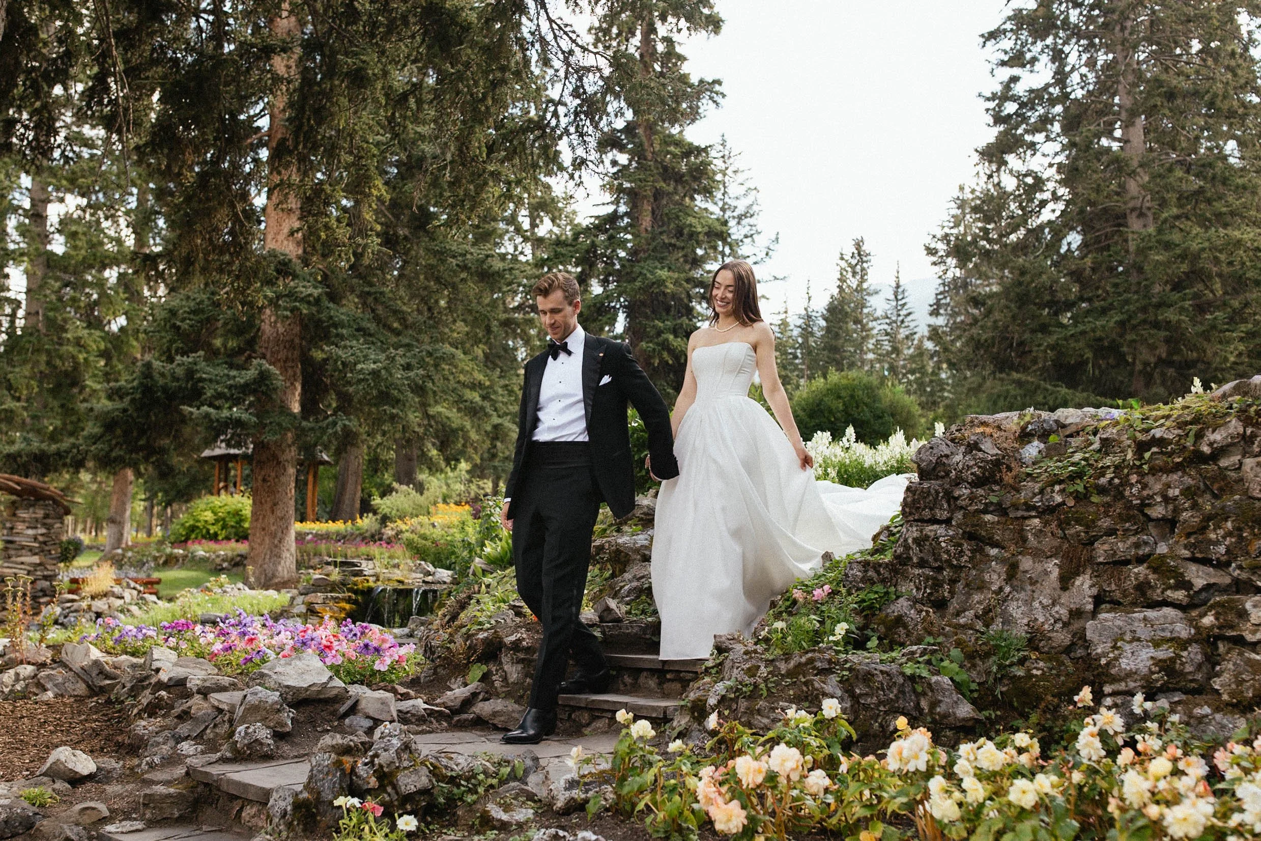 Groom in black tuxedo leads bride in white corset dress down stone steps surrounded by flowers through Cascade Gardens on their wedding day in Banff