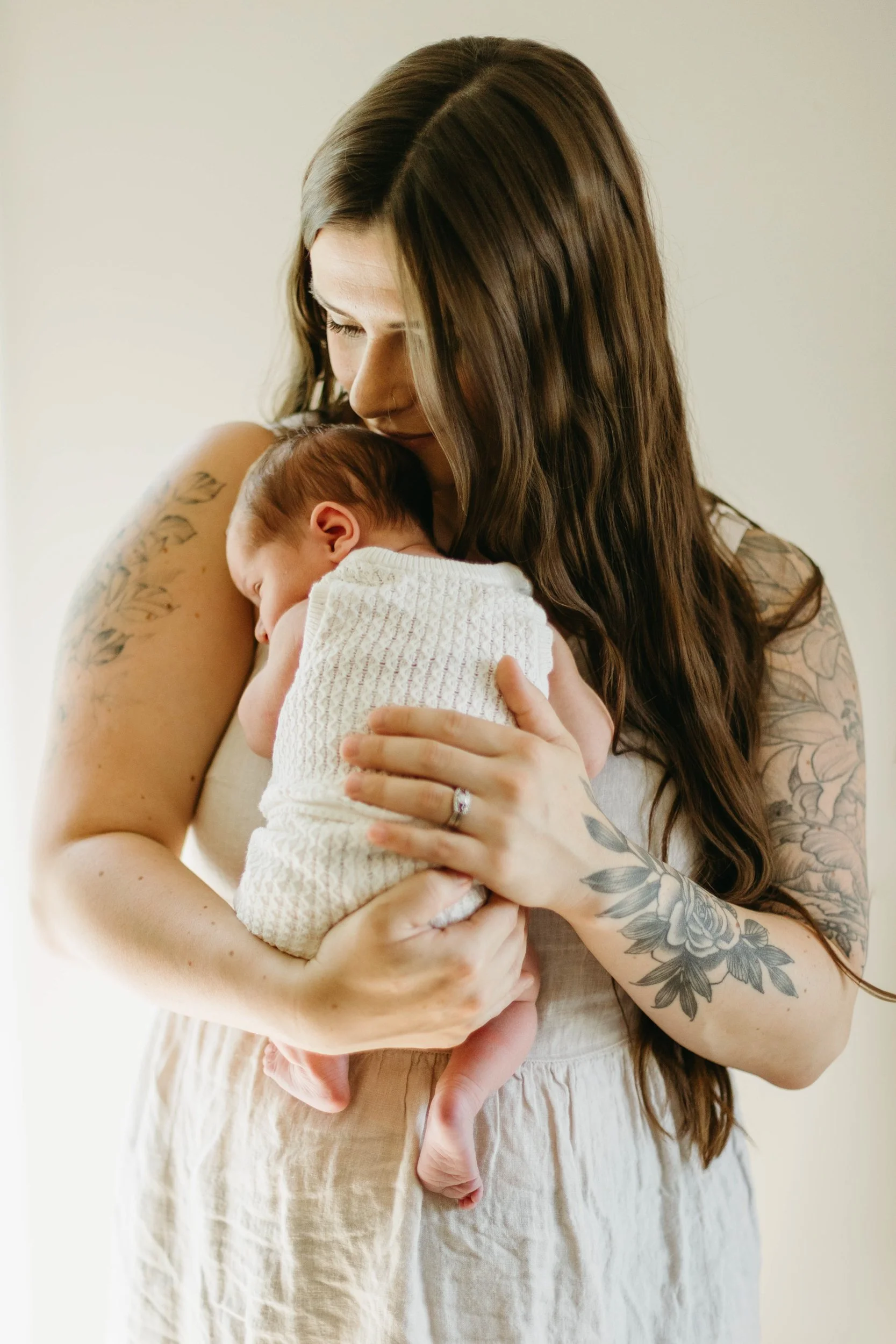 A woman with tattoos and long brown hair cuddles a newborn baby wrapped in a white blanket. She wears a beige dress and shows a wedding ring on her finger.