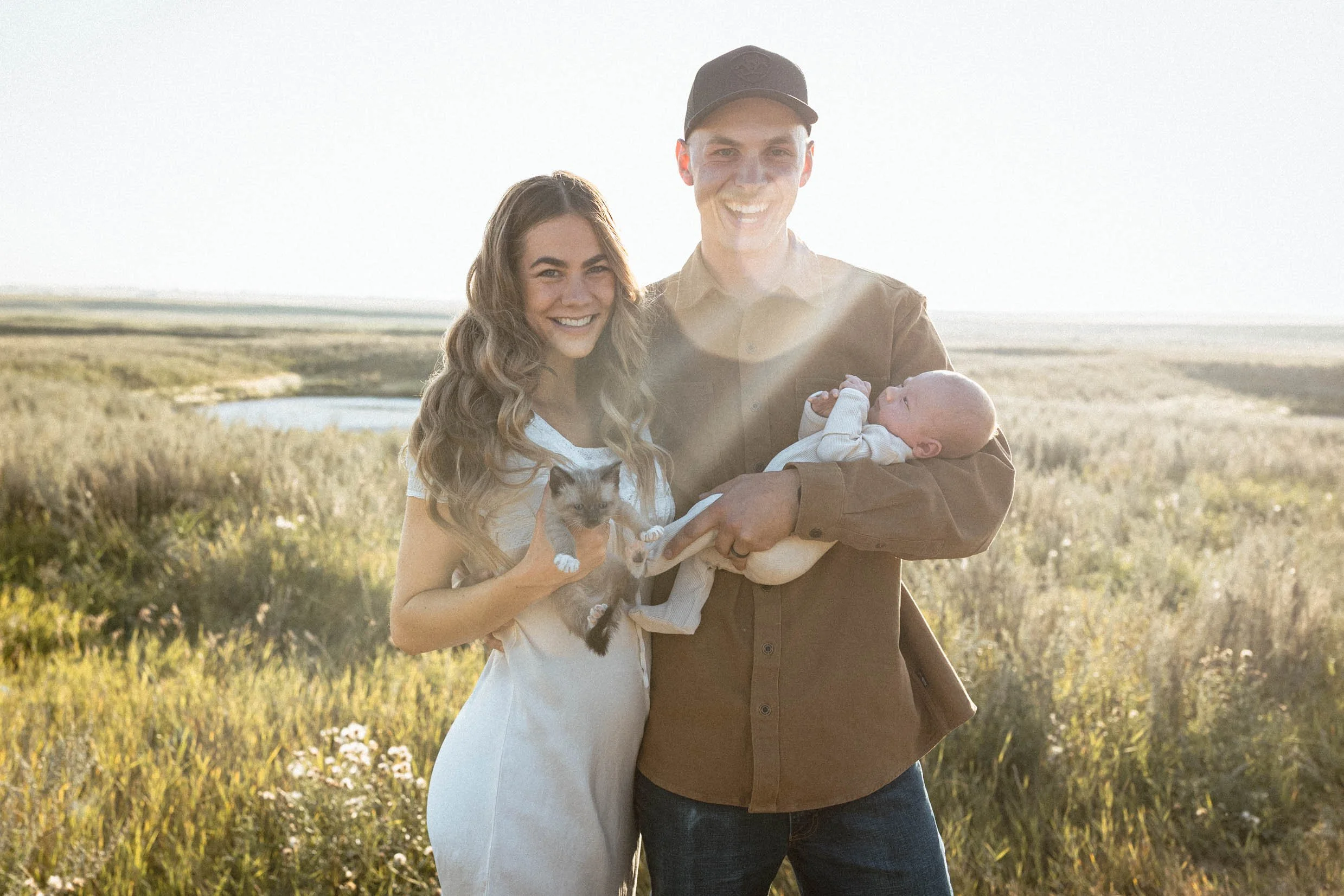 35mm candid film photo of couple smiling in golden sunlight holding their newborn baby and kitten