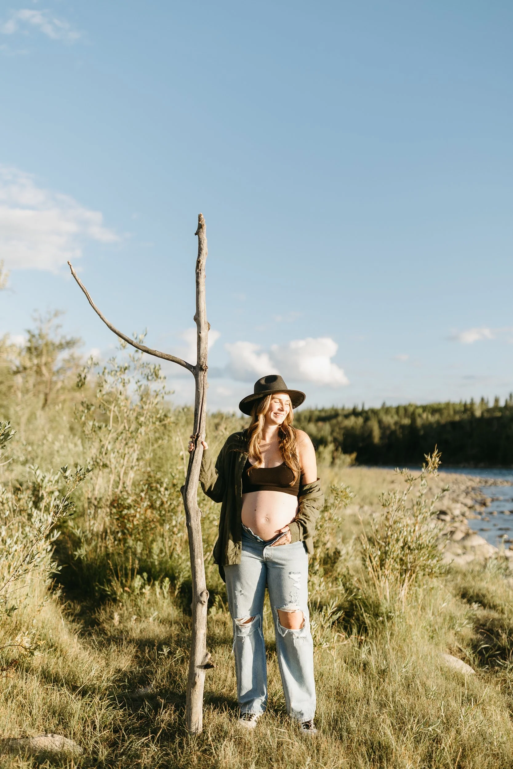 A pregnant woman in a black bralette, ripped jeans, and an olive green jacket standing on a grassy riverside, holding a tall, leafless tree branch, wearing a wide-brimmed hat and smiling in sunlight.