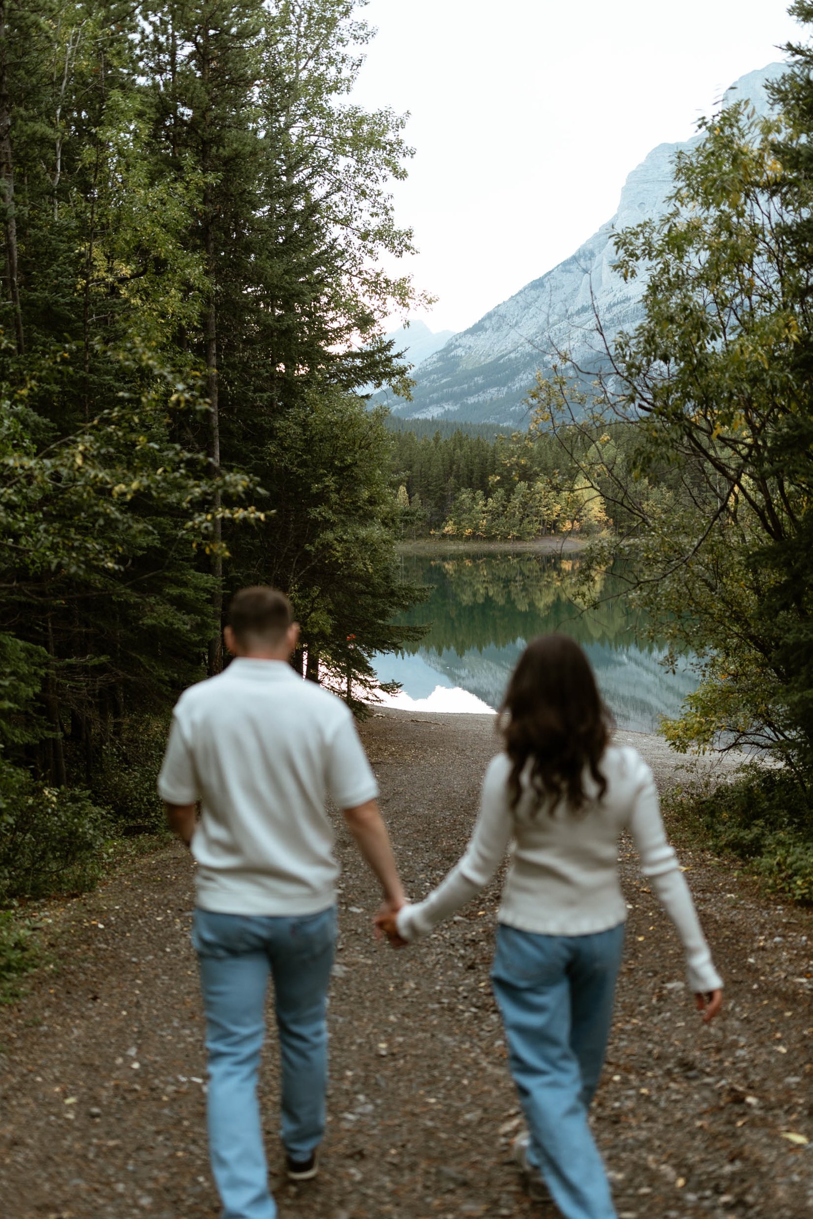 A couple walking hand in hand on a forest trail near a lake, surrounded by trees and mountains in the background.