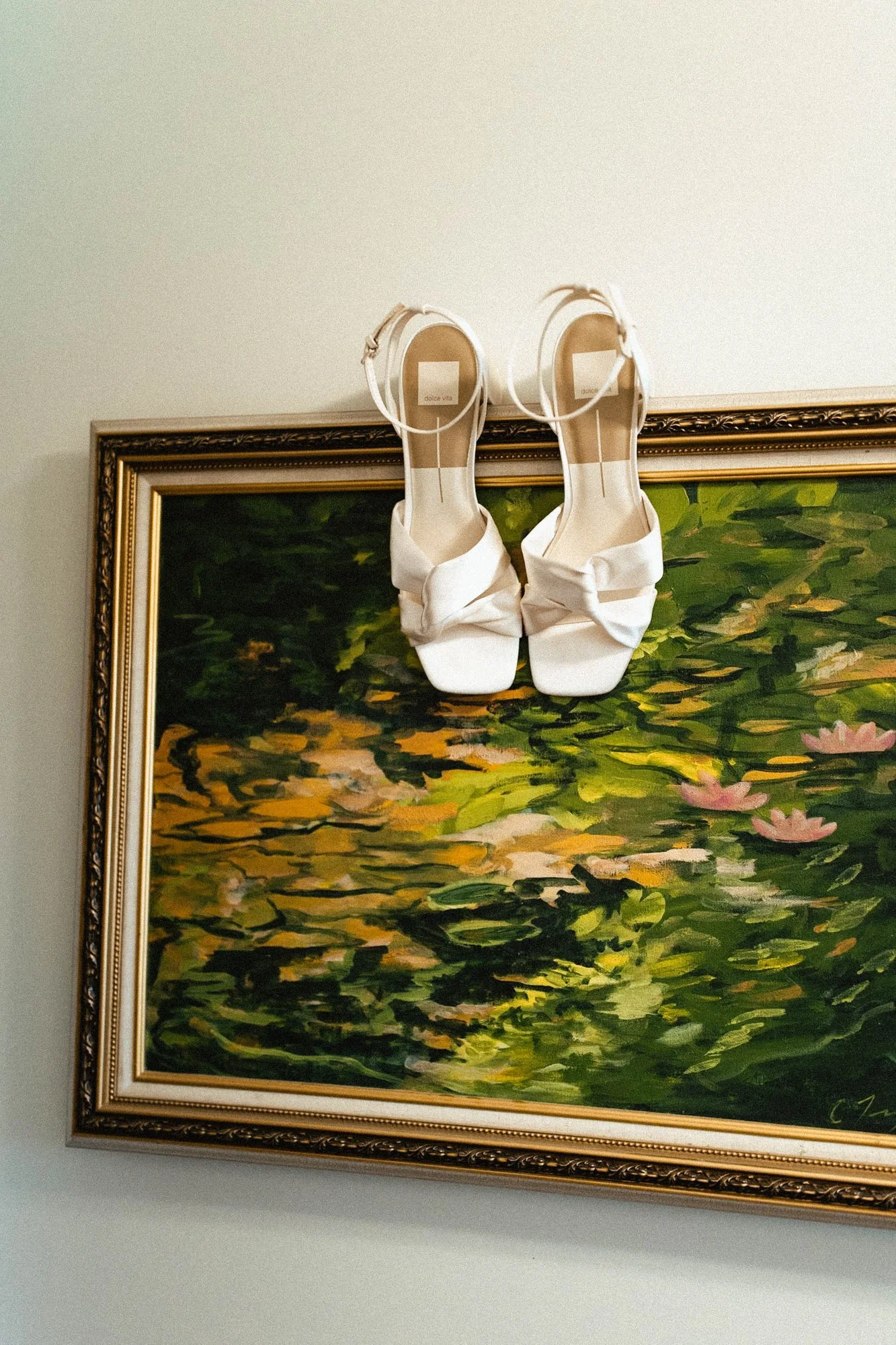 A pair of white high heels with bow accents hanging above a framed landscape painting of water lilies on a pond.