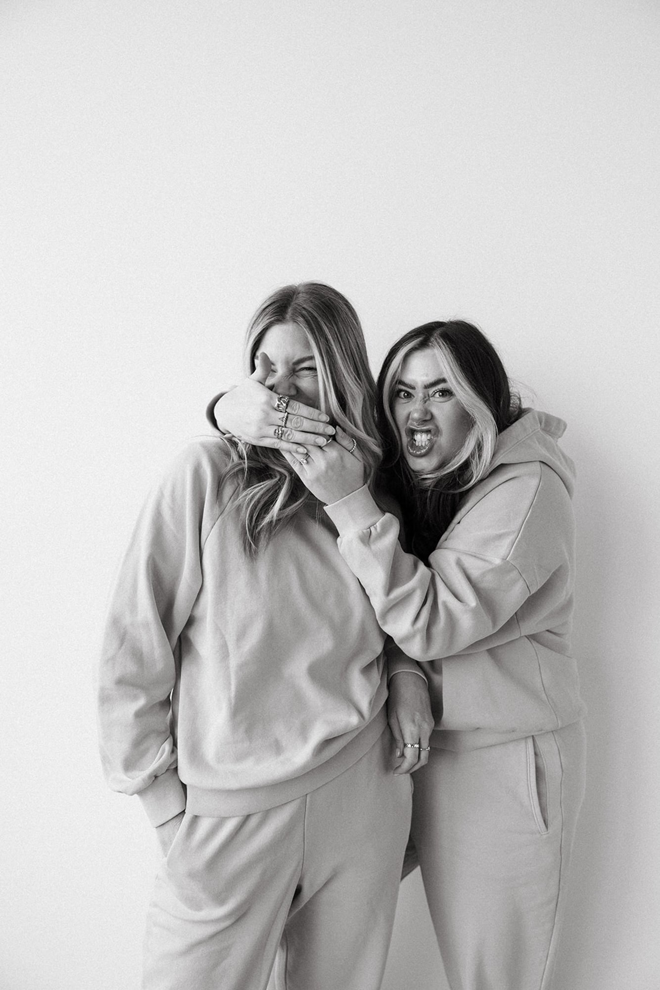 Two women wearing sweatshirts and sweatpants making playful faces and gestures against a plain white background.