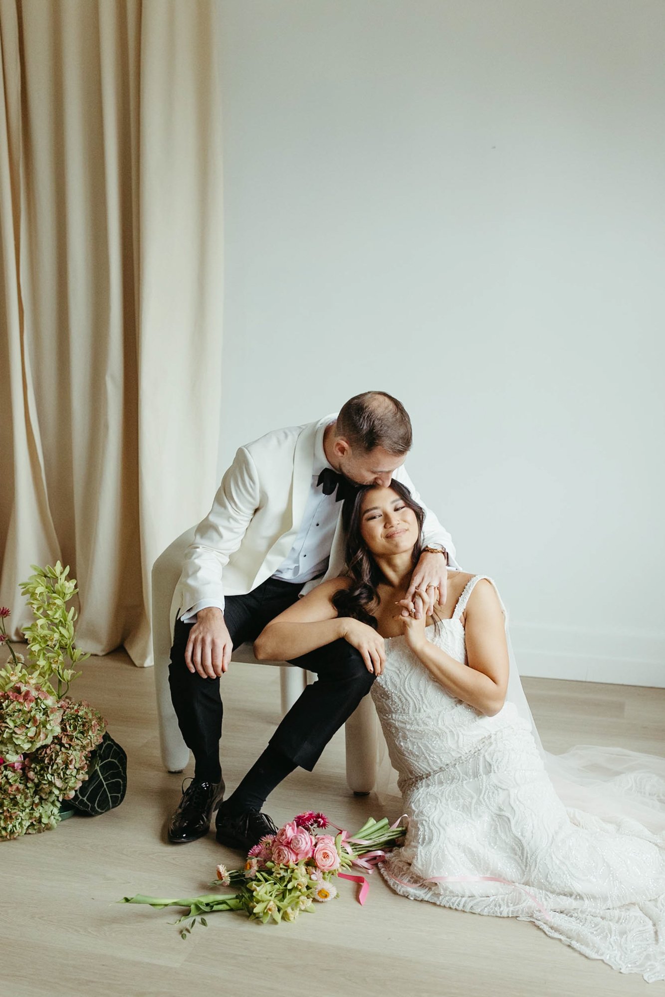A bride and groom pose together in a modern minimalistic setting, with the groom wearing a white tuxedo jacket and the bride in a lace wedding dress, surrounded by bouquets of flowers.