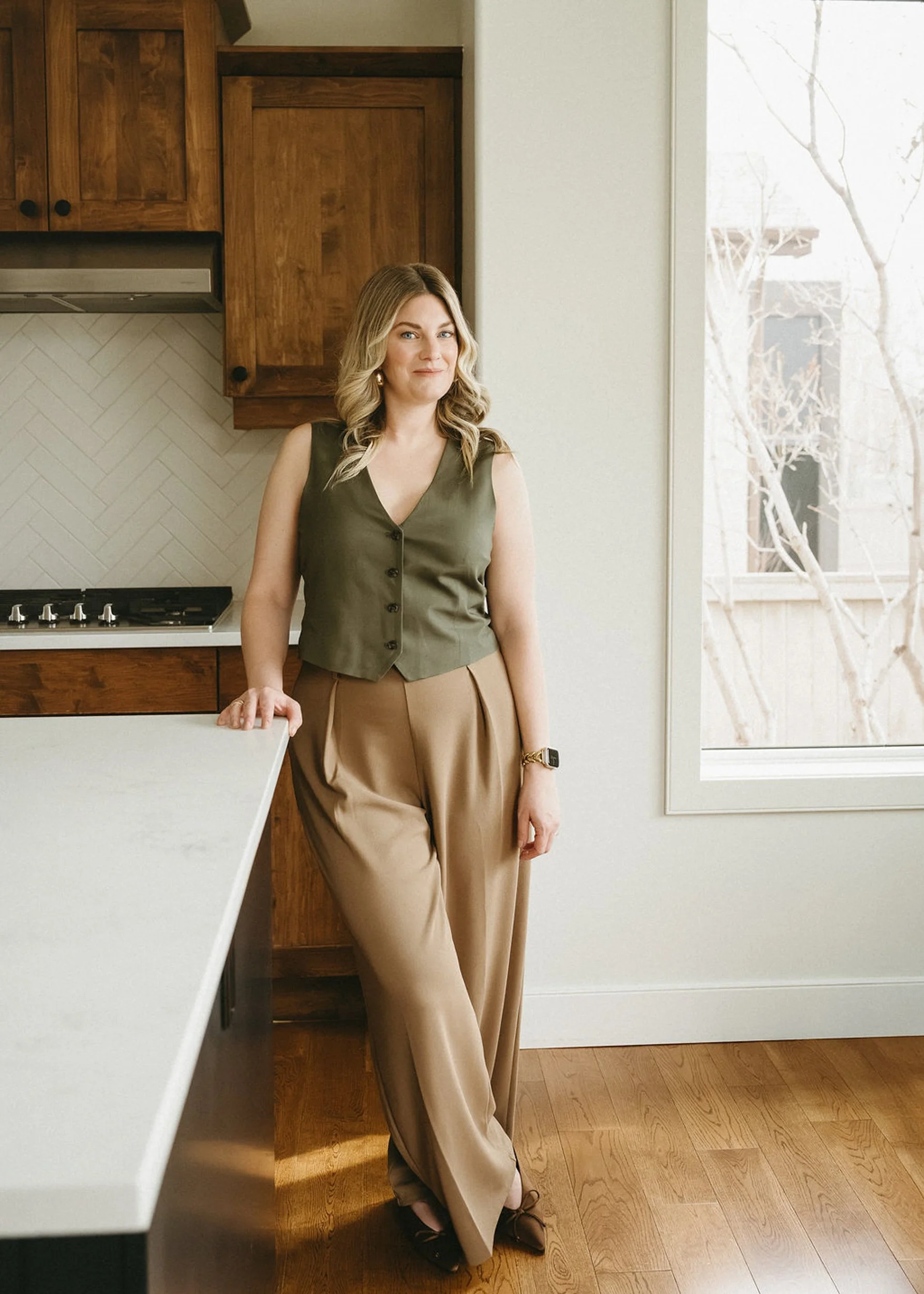 A woman standing in a kitchen near a large window with bare trees outside. She is wearing a sleeveless olive green vest, beige wide-leg pants, and black shoes. She has blonde wavy hair, a watch on her wrist, and is smiling softly.