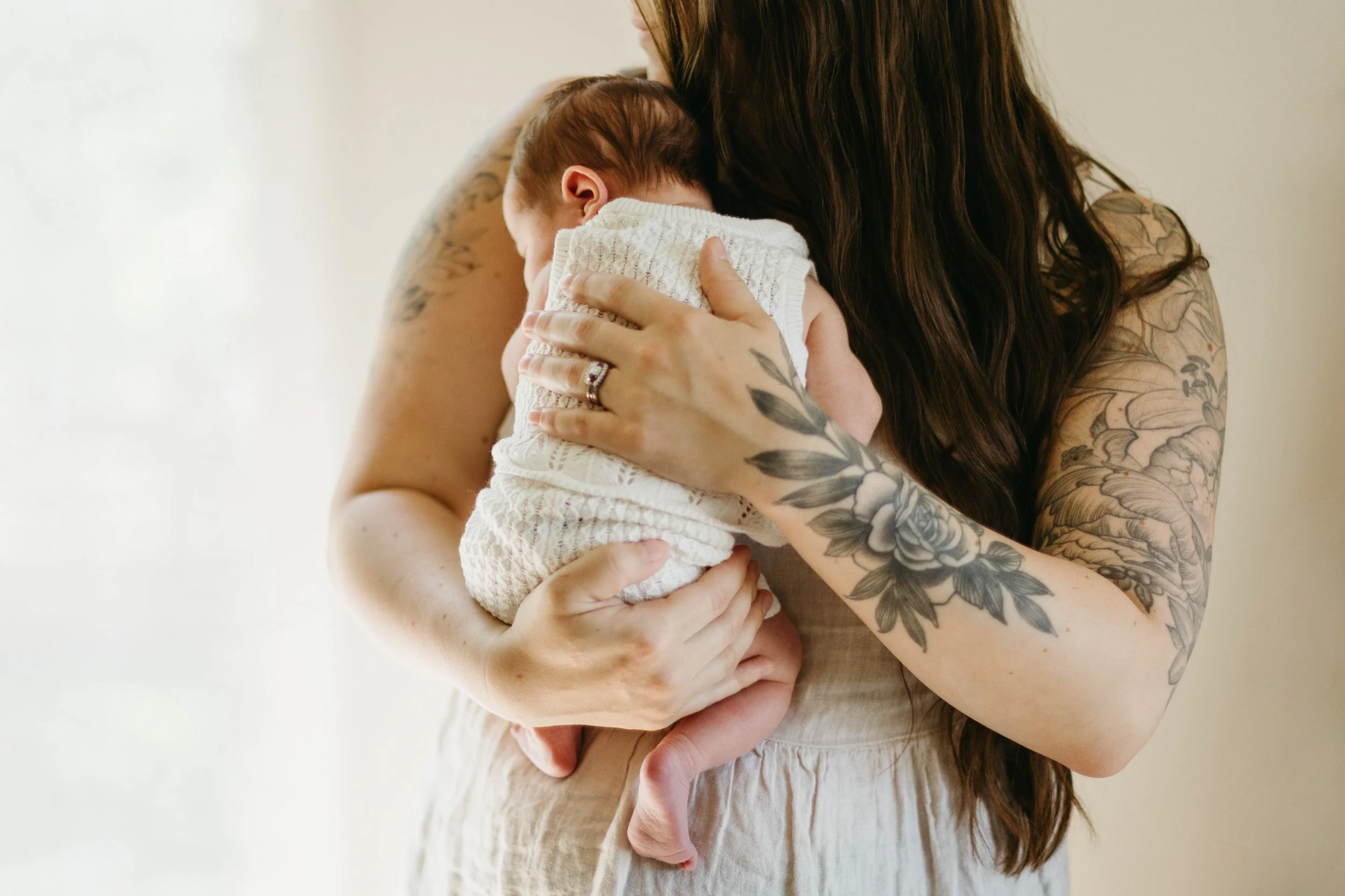 A woman with long dark hair and tattoos on her arms holding a sleeping baby against her chest. The woman is gently cradling the baby in her arms.