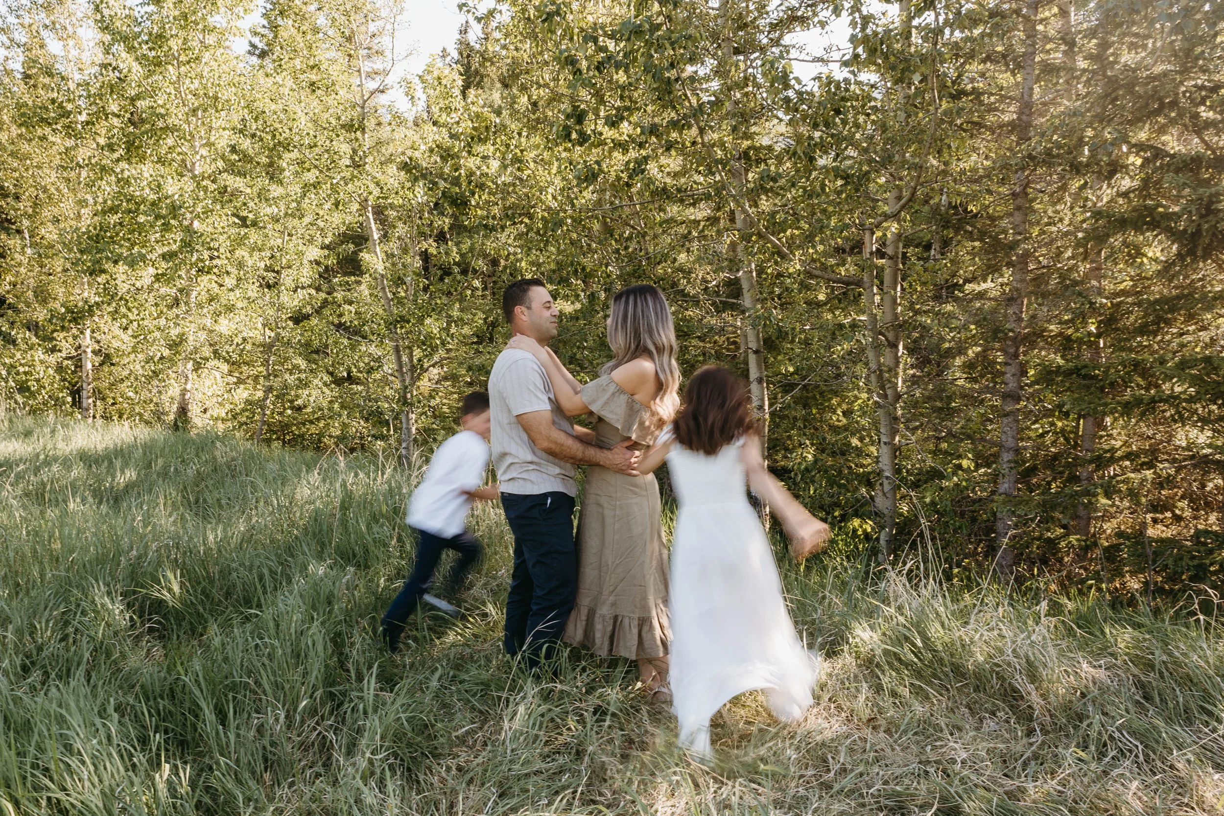 A family of five enjoying time outdoors in a green, wooded area with tall grass and trees, during what appears to be late afternoon or early evening.