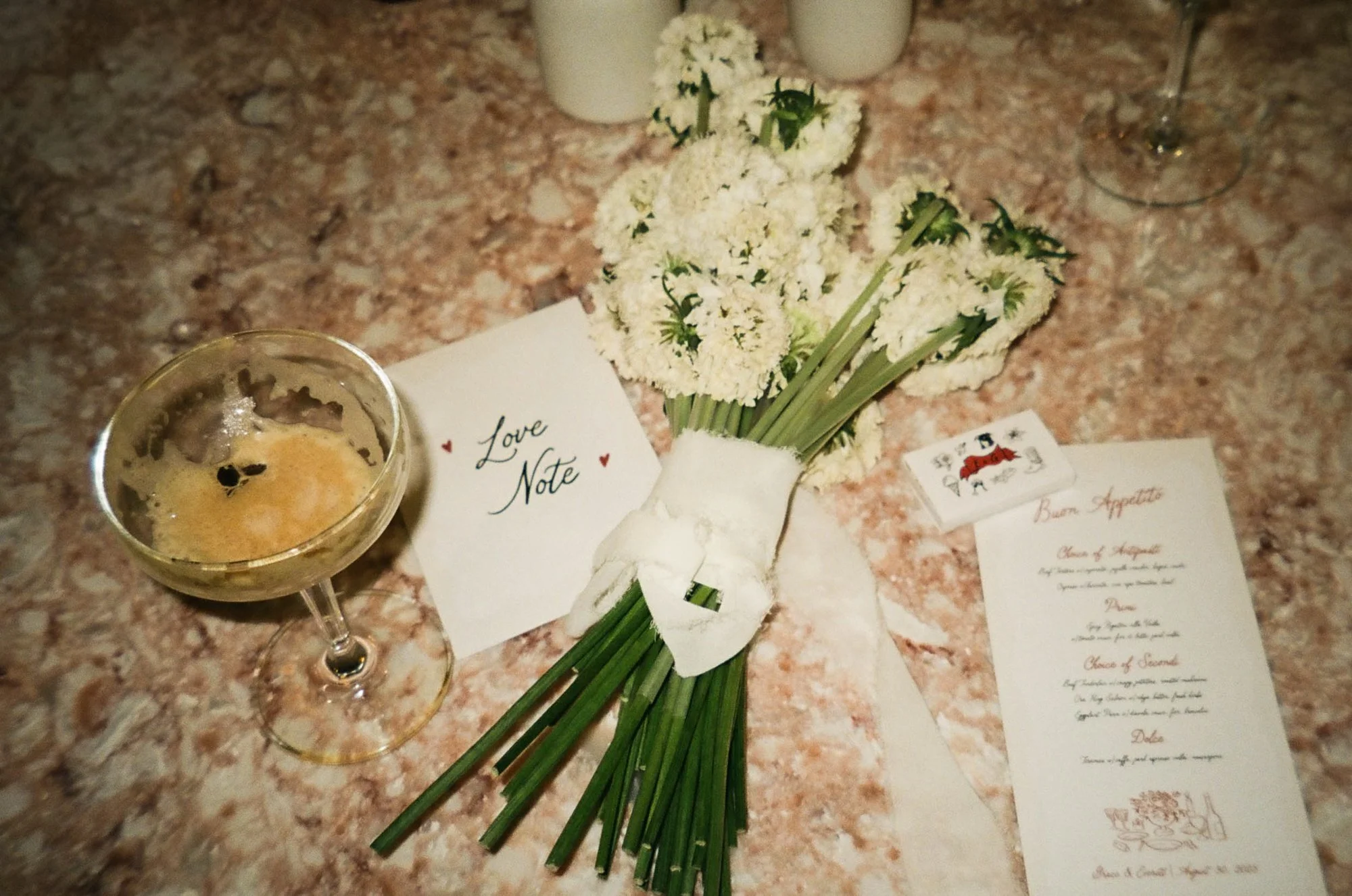 A table with a bouquet of white flowers, a love note card, a candle in a glass holder, a small sticker with a dog illustration, and a printed menu with Italian dishes.