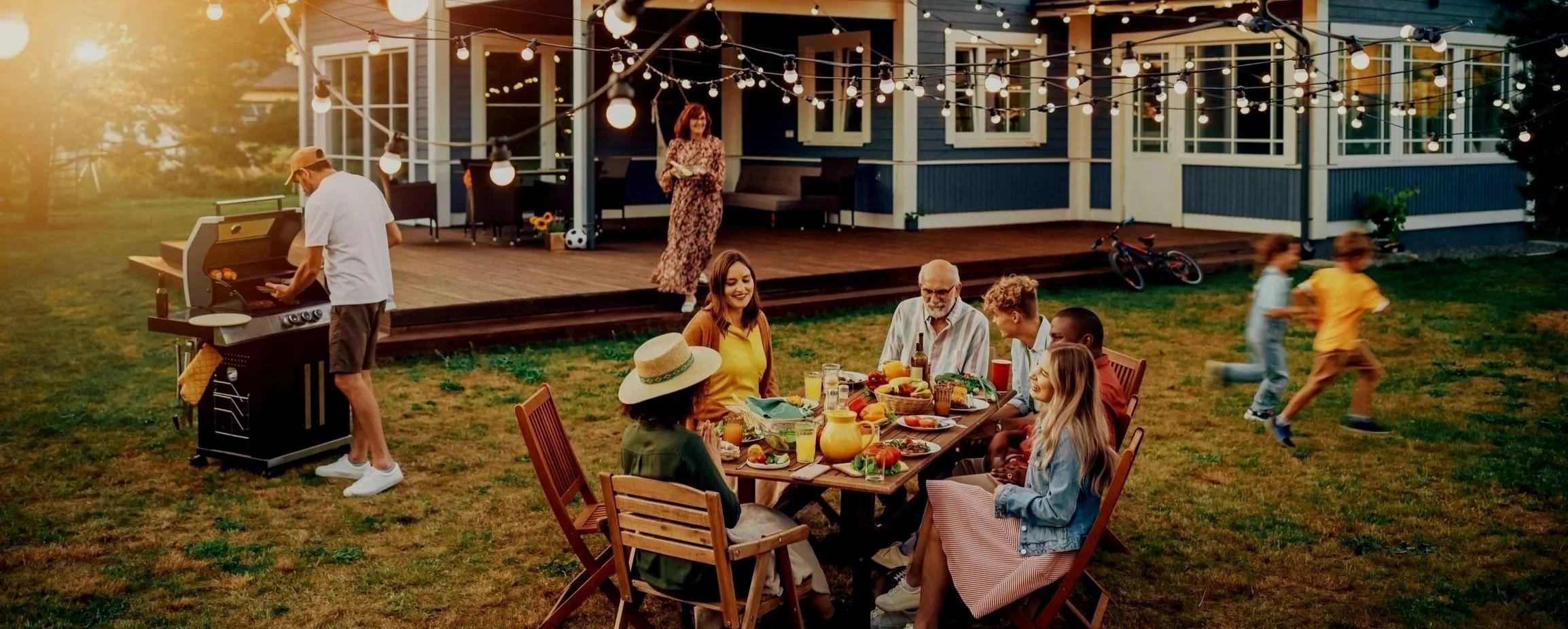 Family and friends enjoy outdoor dinner party in backyard with string lights, children running, and a grill chef cooking. House with large windows in background.