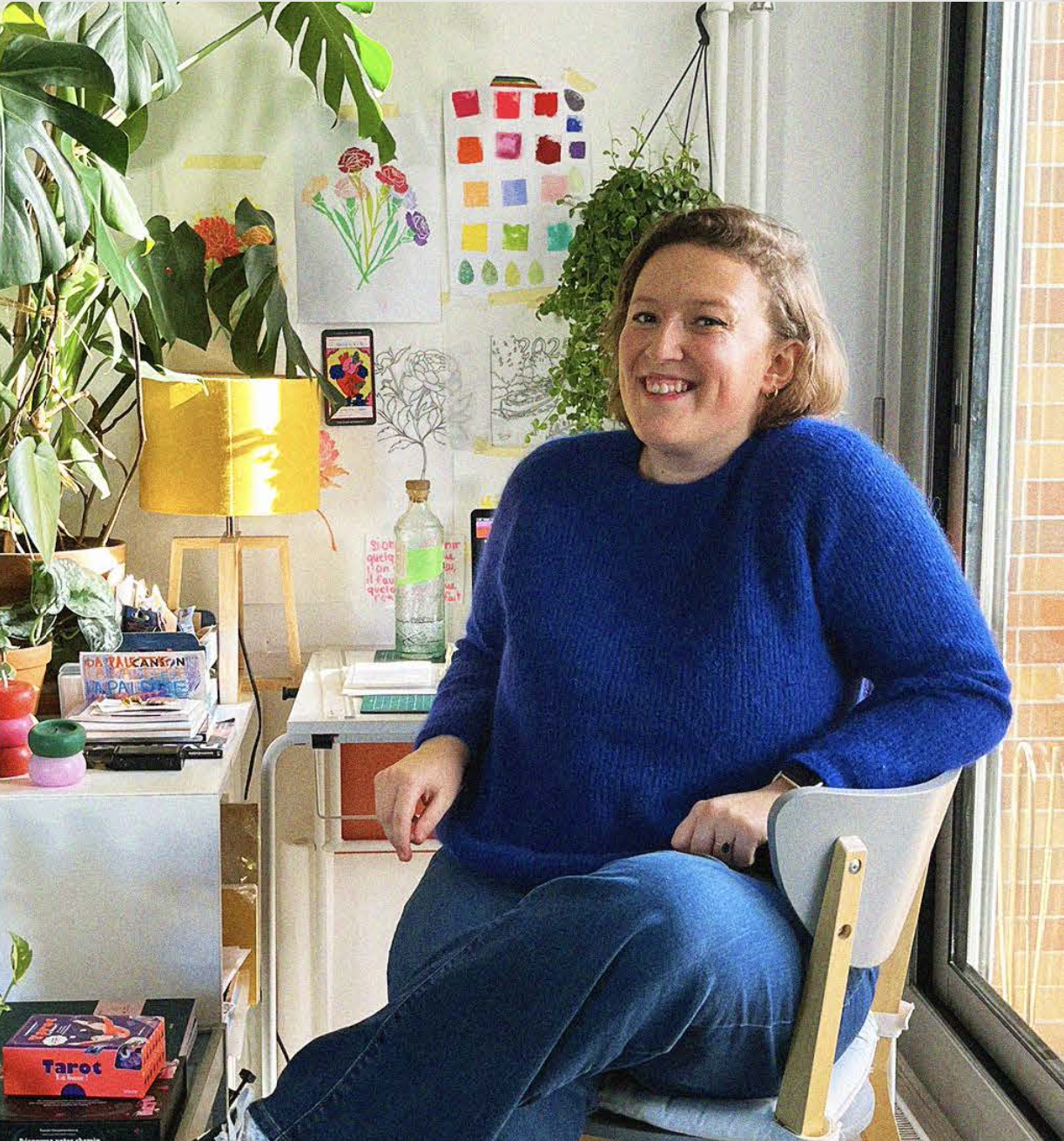 Une femme souriante assise dans une pièce avec des plantes, un lampadaire jaune, et des dessins et peintures colorés au mur, près d'une fenêtre.