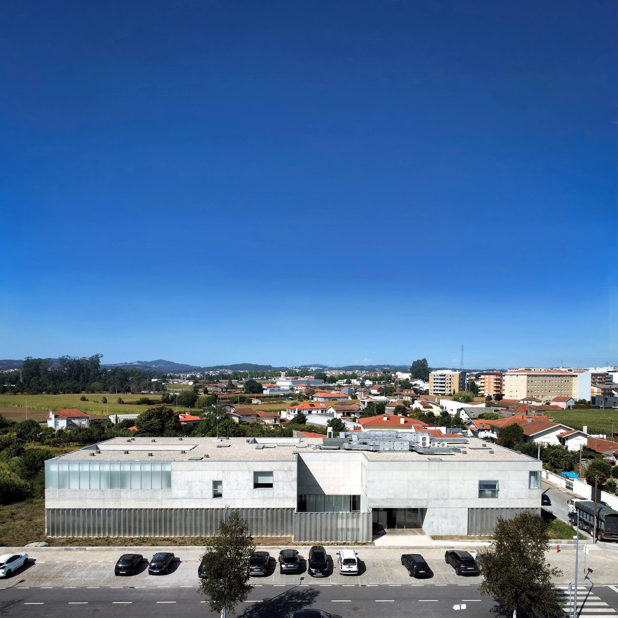 Foto de um edifício moderno de cor cinza em frente a uma rua com carros estacionados, ao fundo, uma cidade com casas e prédios e céu azul claro.