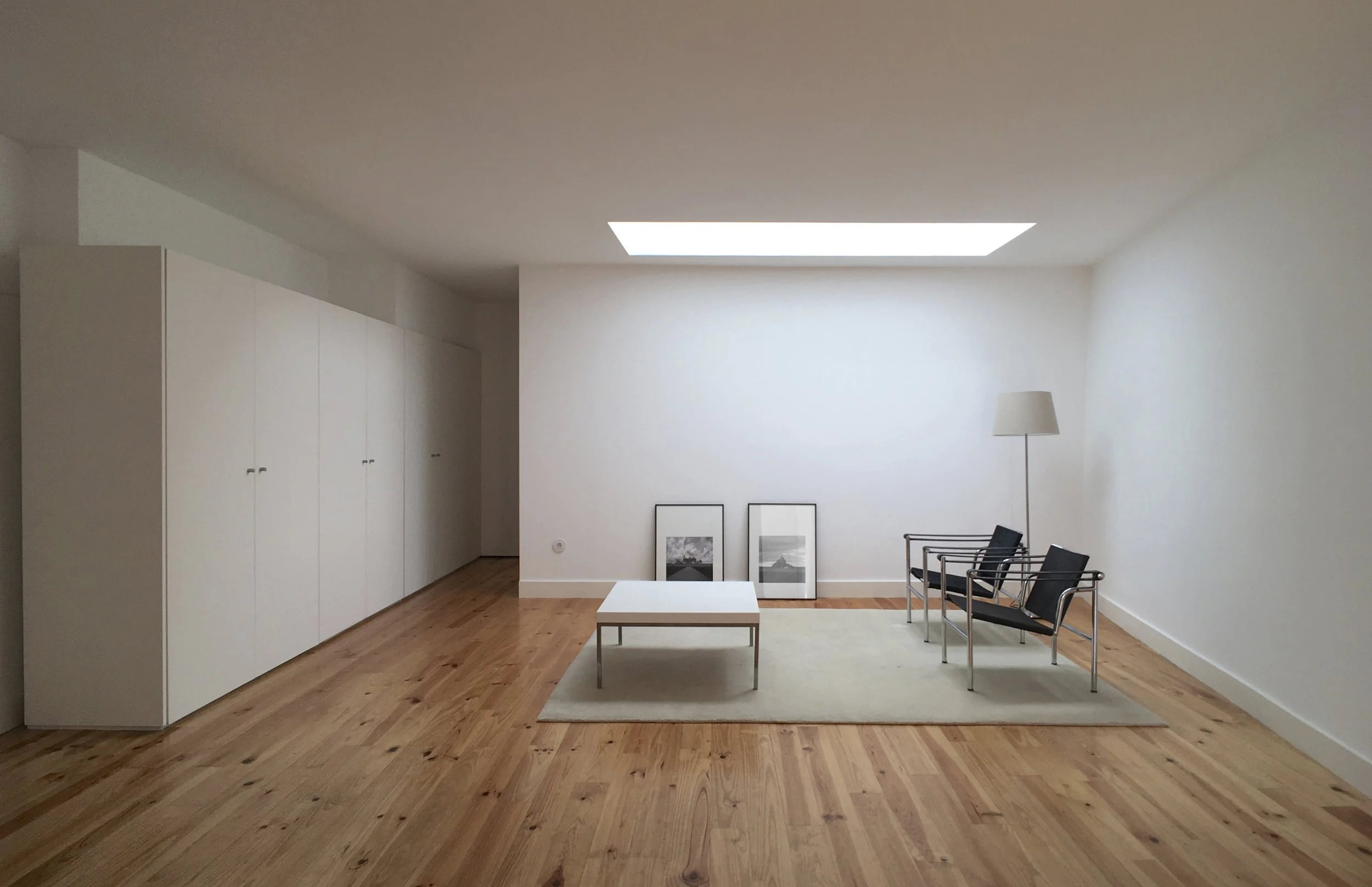 Sala de estar minimalista com chão de madeira, mesa branca, dois quadros com fotos preto e branco apoiados na parede, dois cadeiras pretas com estrutura metálica, par de abajures e um tapete claro.