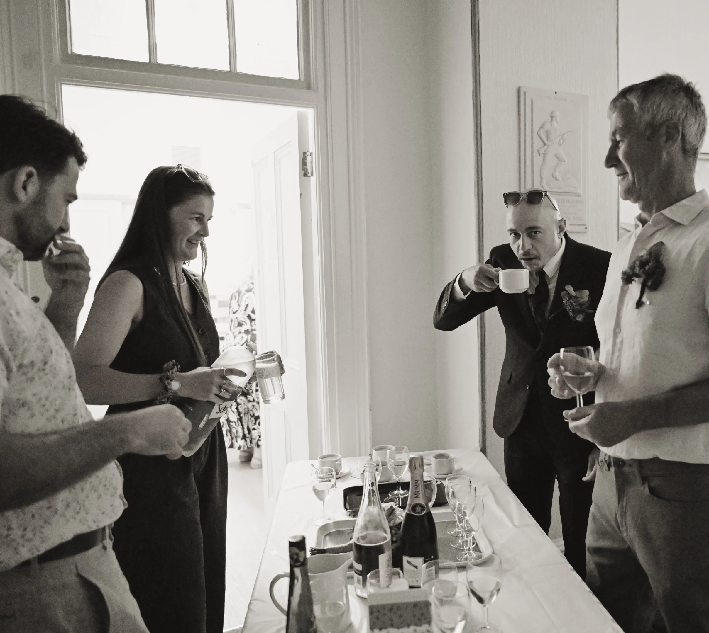A black and white photo of five people gathered around a table with drinks, at a social event or gathering, indoors near a doorway.