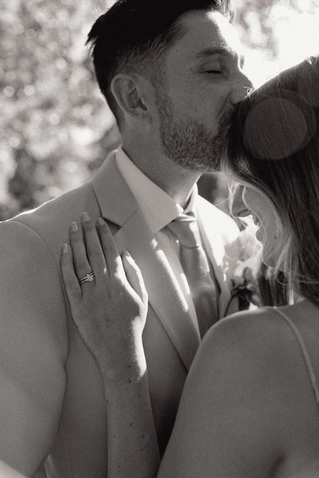 A black-and-white photo of a couple kissing, with the woman's hand showing a wedding ring, and the man dressed in a suit.