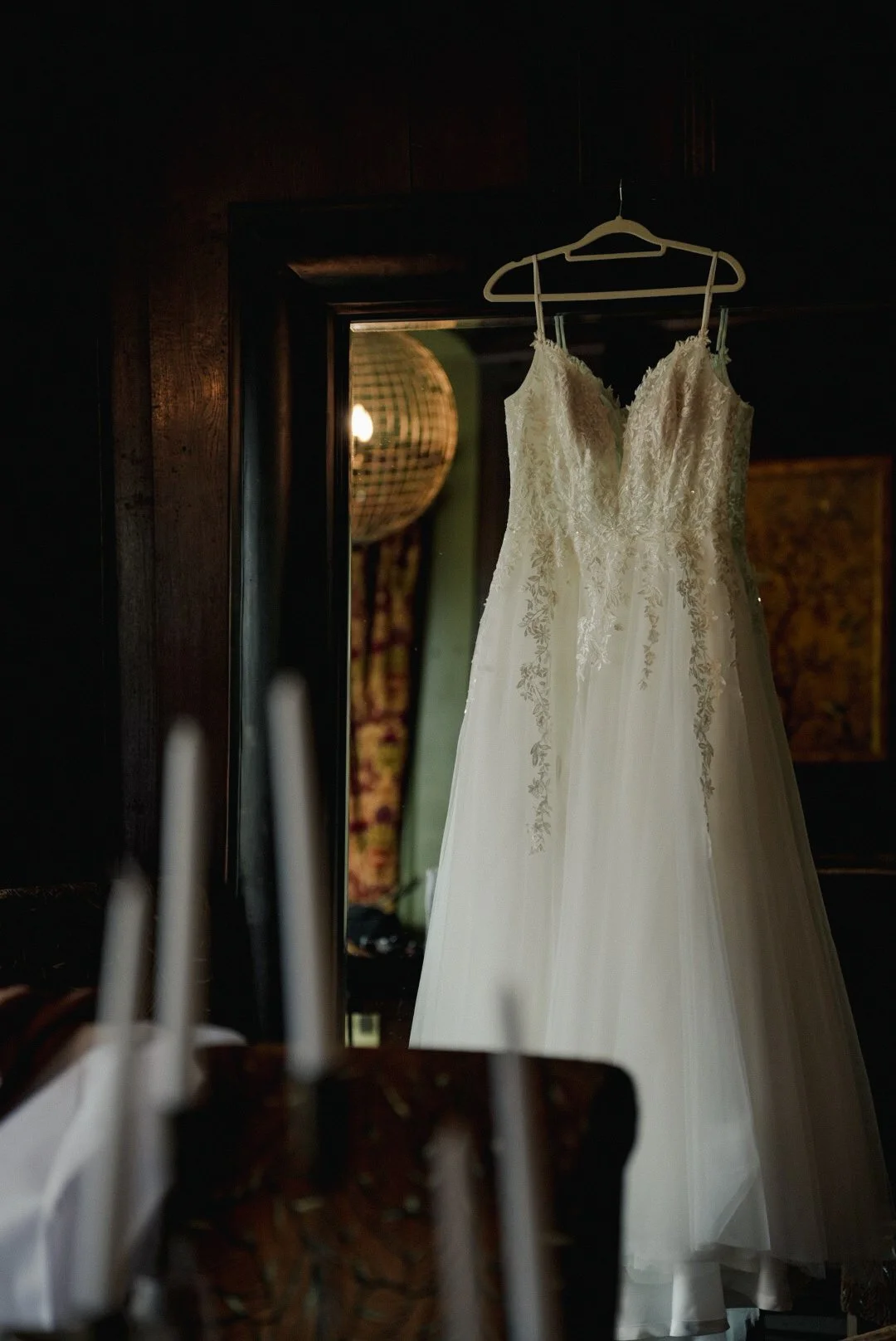 A white wedding dress hanging on a hanger in front of a mirror in a dimly lit room.