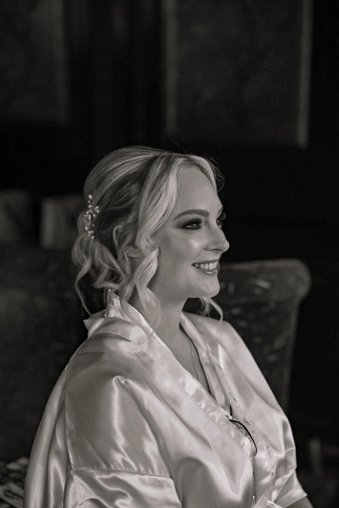 Black and white photo of a smiling woman with wavy blonde hair, wearing a satin robe and decorative hairpiece, sitting indoors against a dark wooden background.
