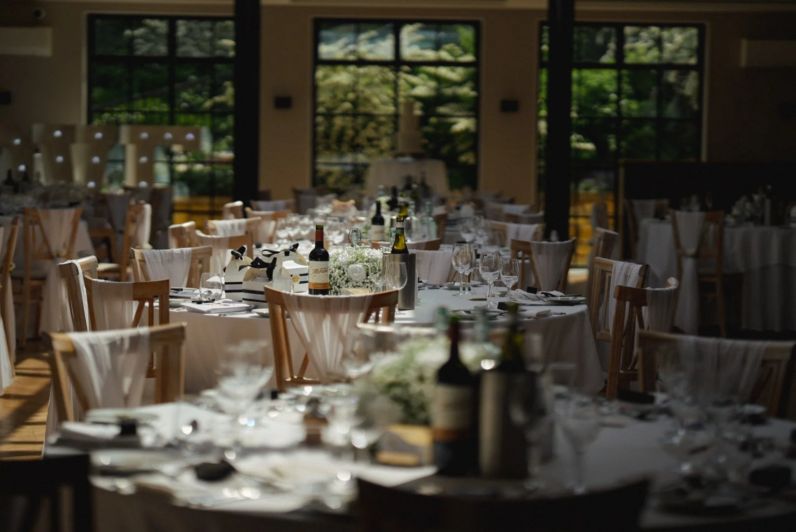 A banquet hall set up for a formal event with round tables, white tablecloths, wine bottles, and elegant tableware, with large windows showing green trees outside.