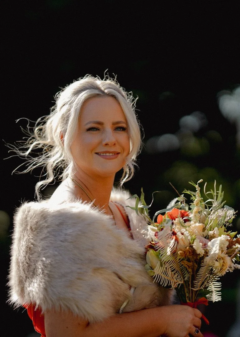 A woman with blond hair holding a bouquet of flowers, smiling, outdoors with a dark, blurred background.