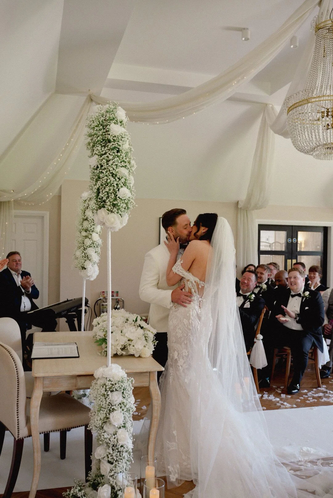 A bride and groom kiss at their wedding ceremony, surrounded by seated guests in tuxedos and dresses, with floral decorations and a chandelier in the background.