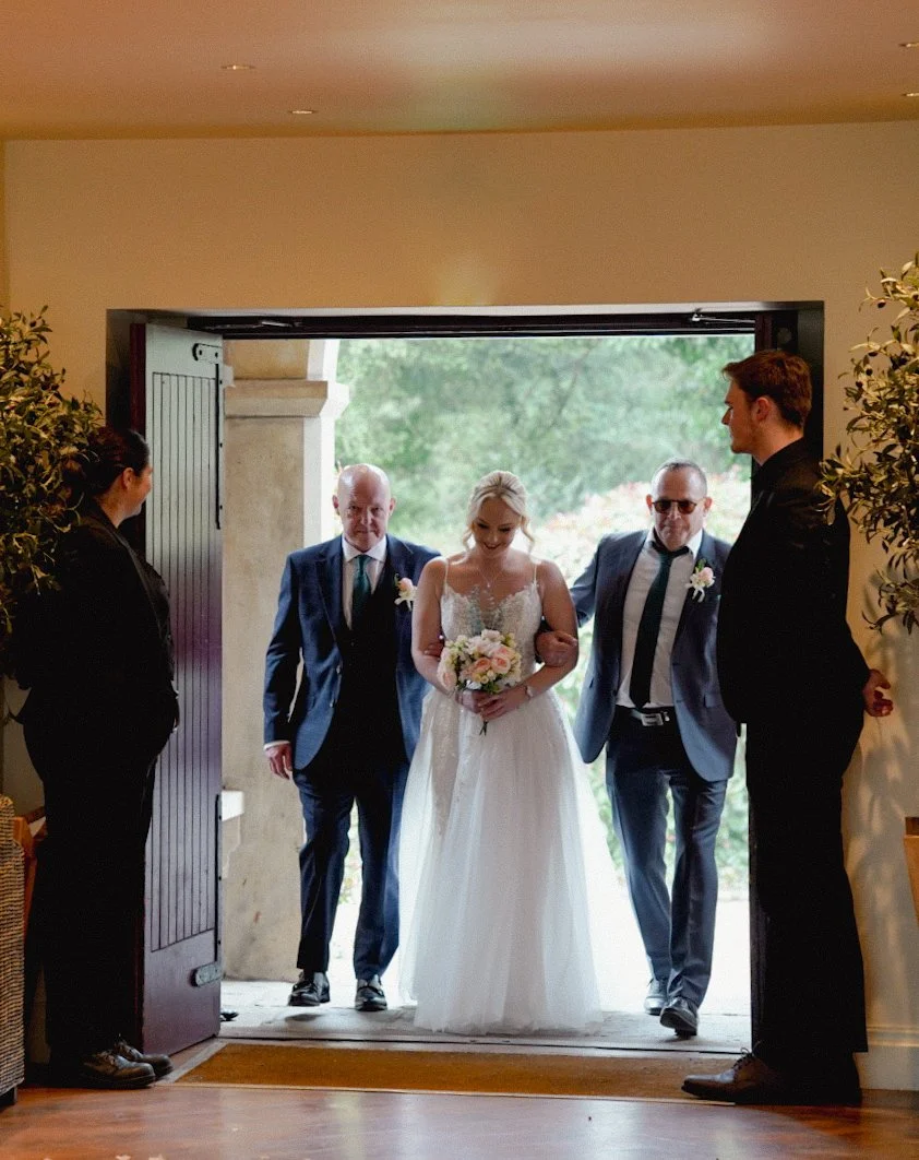 Bride in wedding dress entering with her parents, flanked by two staff members, in a doorway with trees visible outside.