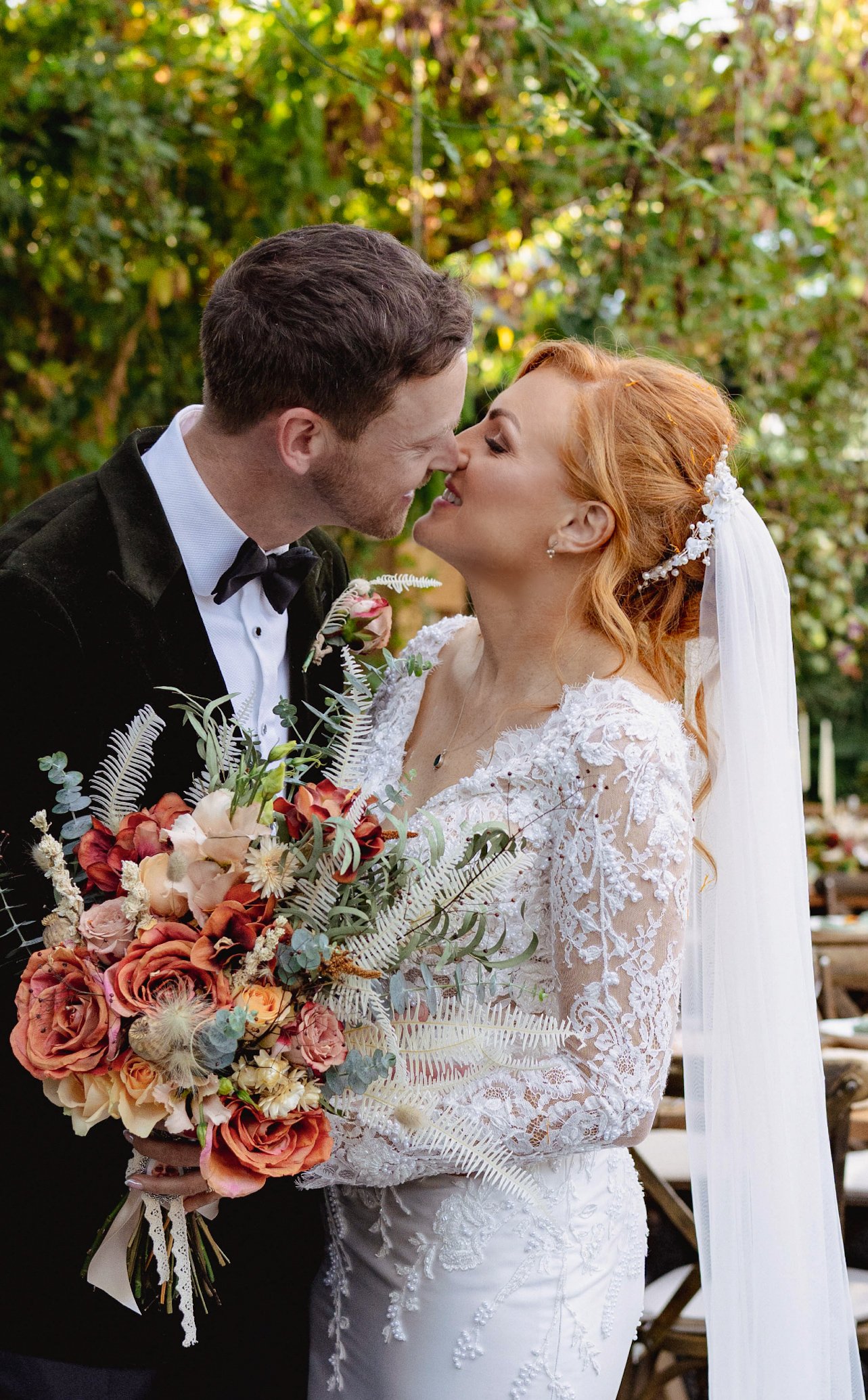 A bride and groom are close together, about to kiss, outdoors during daytime, with the bride holding a large bouquet of flowers.
