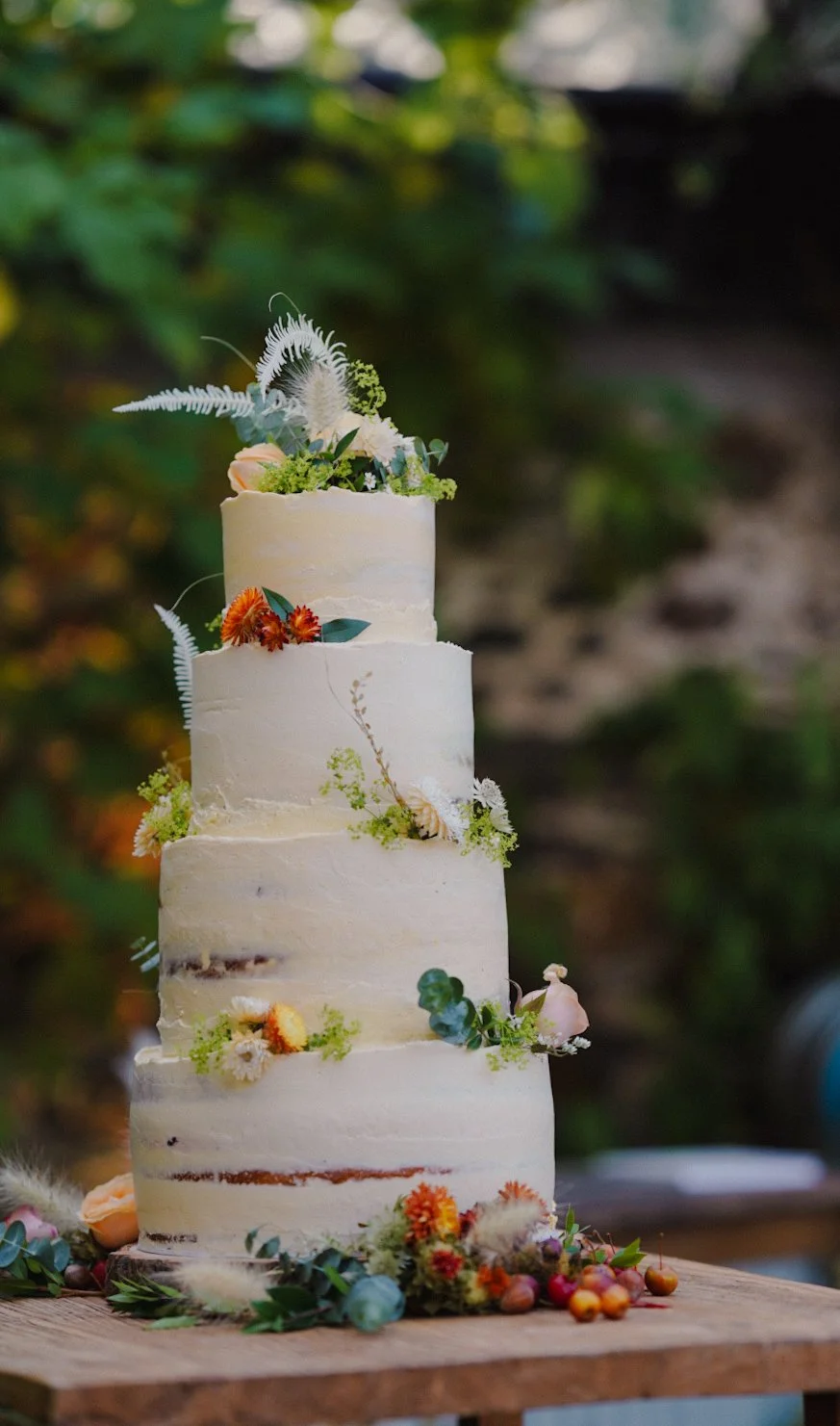 A four-tiered wedding cake decorated with fresh flowers and greenery, set on a wooden table outdoors.