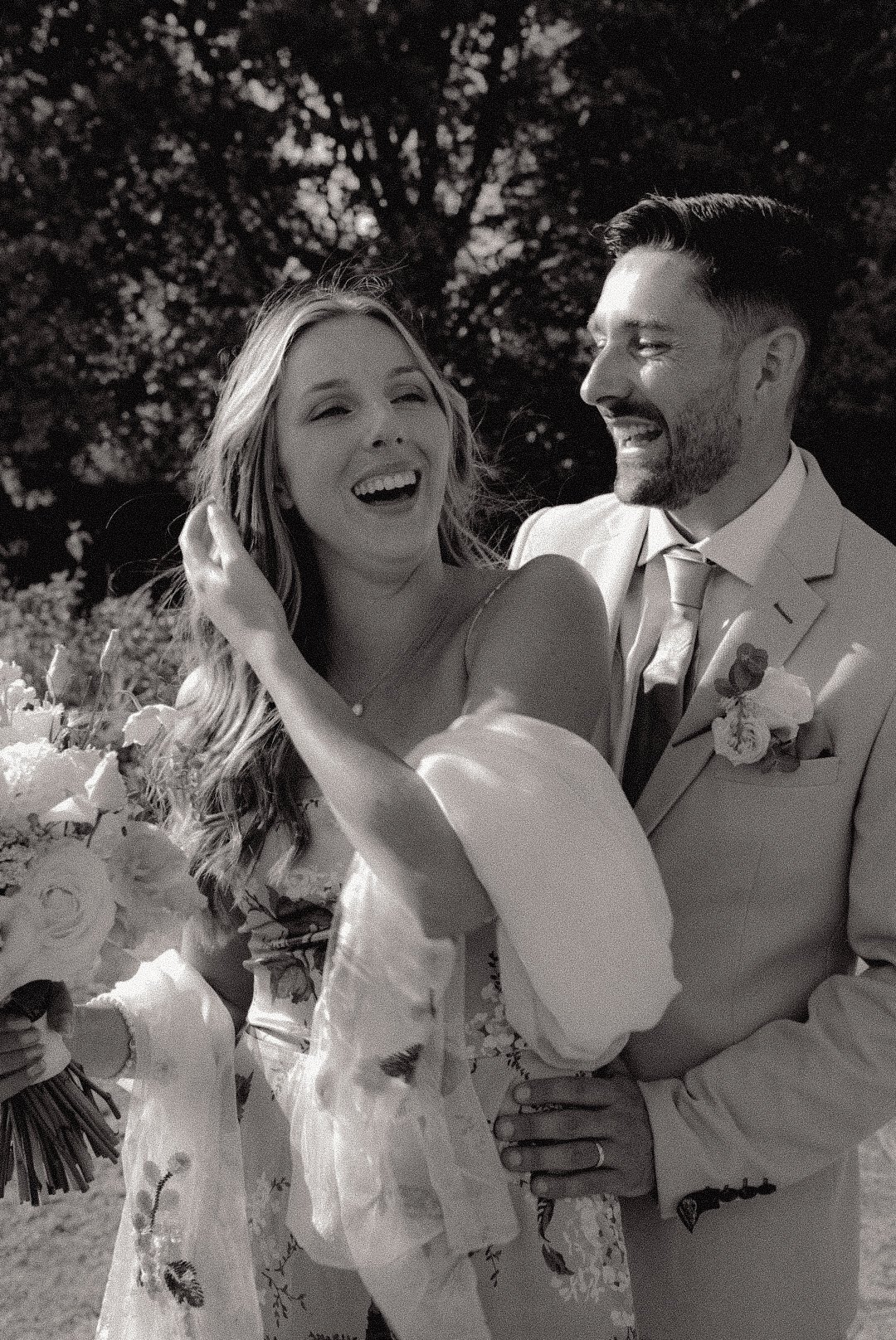 A joyful bride and groom sharing a moment outdoors, dressed in wedding attire, smiling and laughing. The bride is holding a bouquet, and the background features trees.
